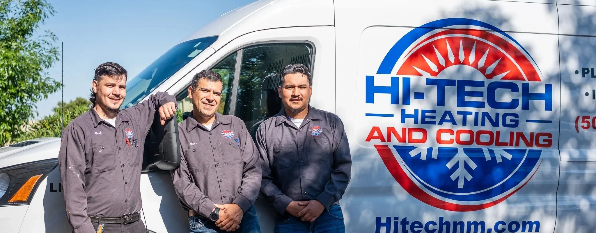 Three technicians in gray work shirts standing in front of a white HVAC service van with the logo of Hitech Heating and Cooling. The man on the left has his arm around the shoulders of the man in the middle. All are smiling and looking at the camera.