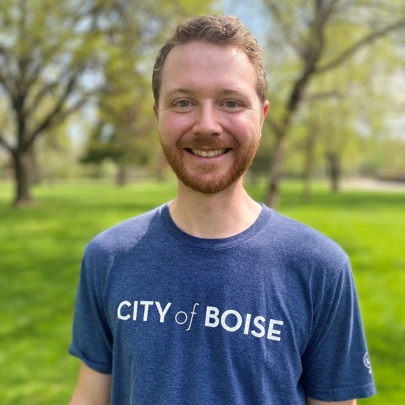 A smiling man with short curly hair and a beard, wearing a blue t-shirt that says 'CITY of BOISE,' standing outdoors in a park with green grass and trees in the background.