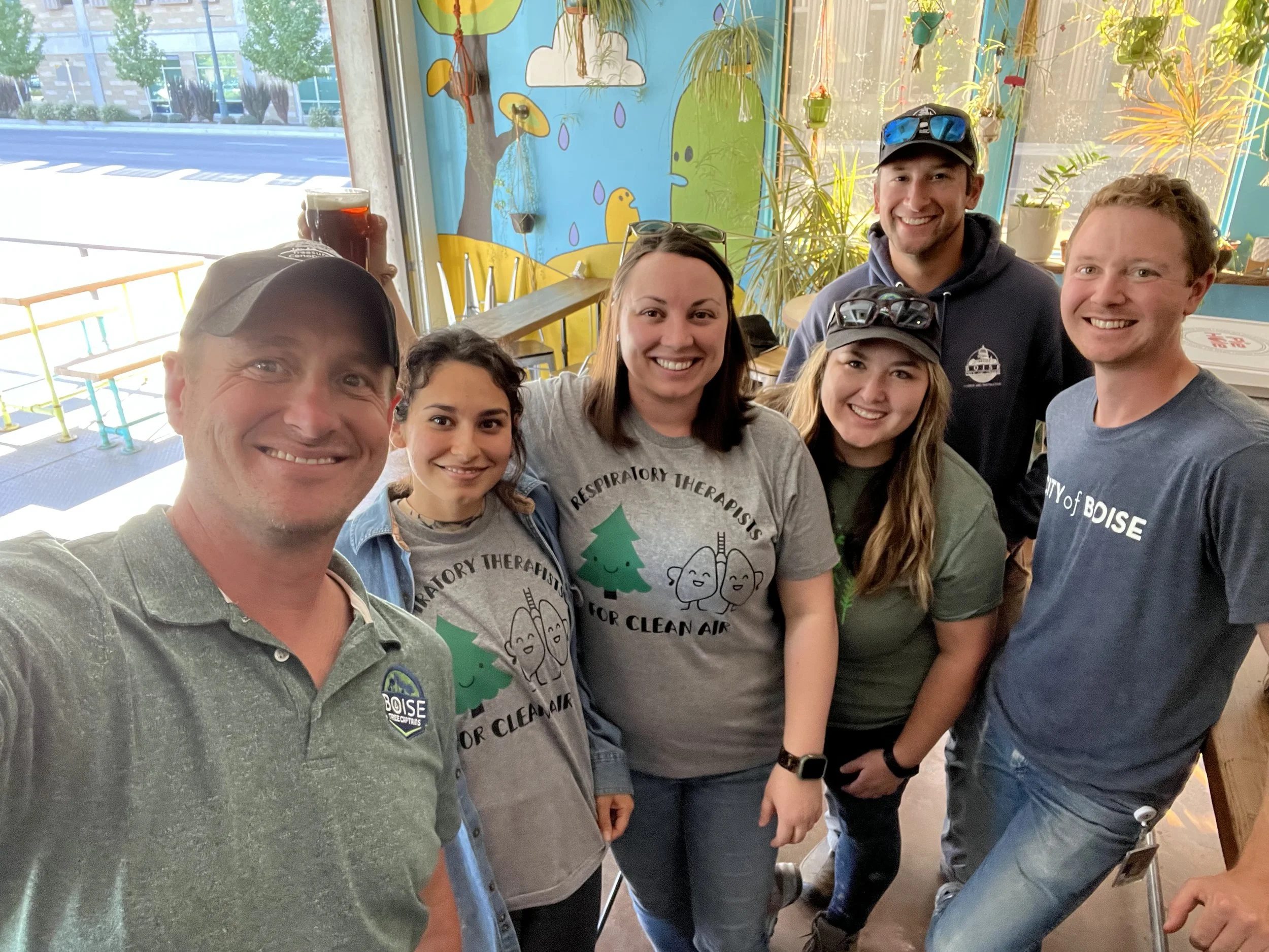 Group of seven smiling people posing together inside a restaurant with colorful blue wall mural and potted plants, some wearing Boise City T-shirts.