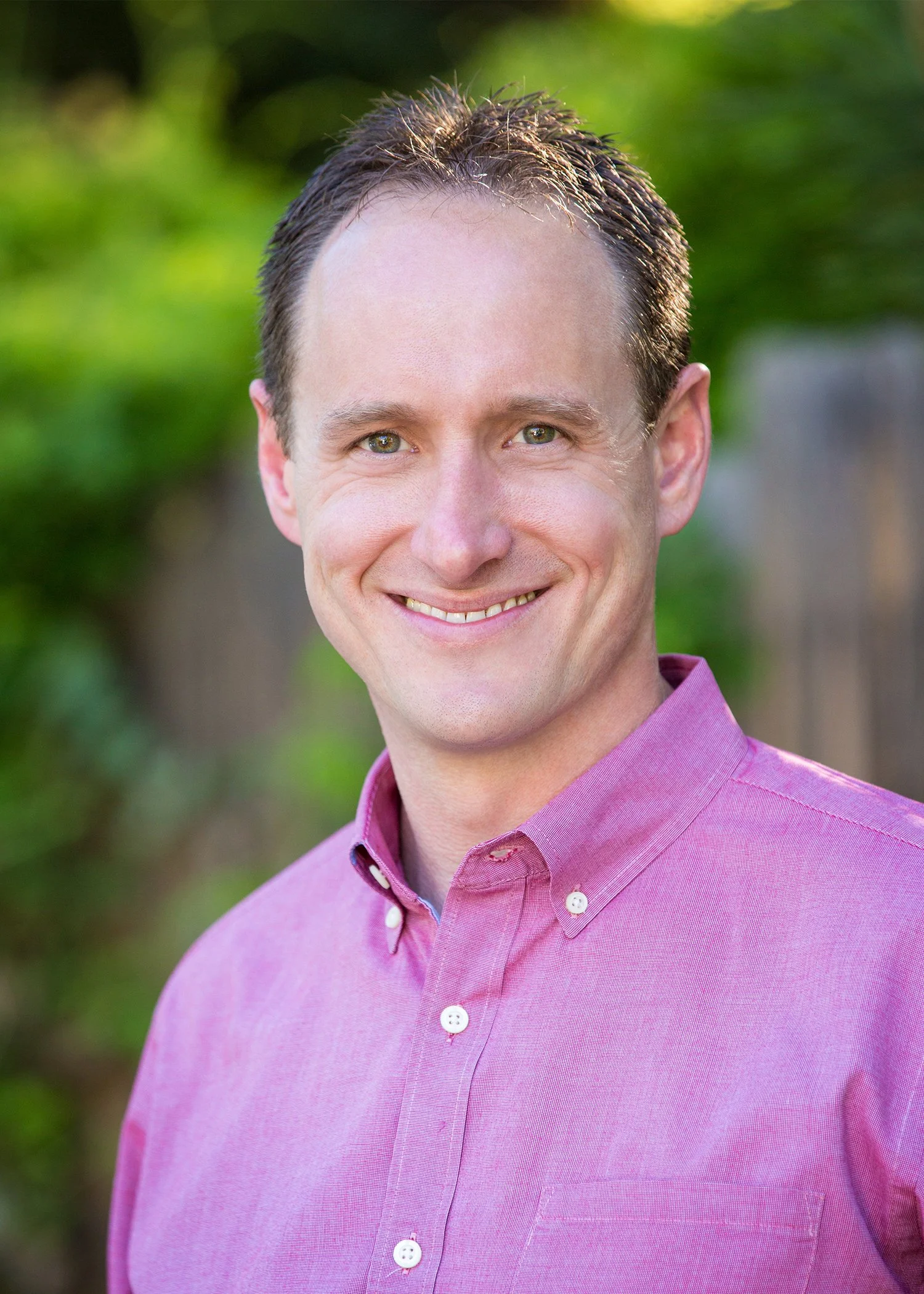 A man smiling outdoors, wearing a pink button-down shirt with a blurred green background.