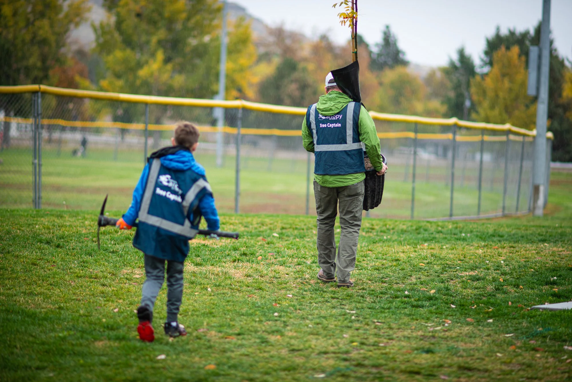 A young boy and an adult walk on a grassy field near a baseball fence. Both are in blue safety vests that say 'Tree Captain' and are holding tools. The scene appears to be at a park during fall.