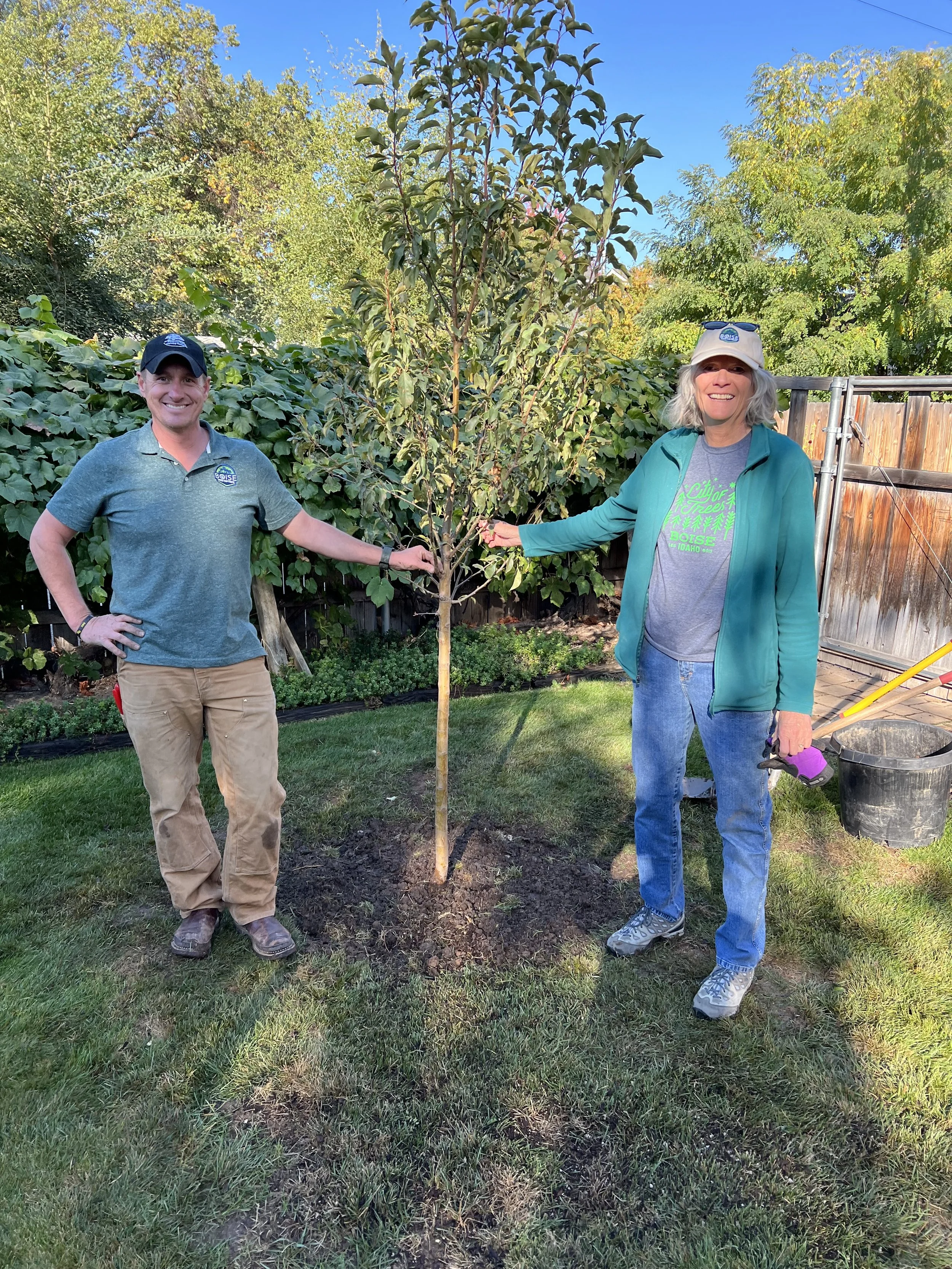 Two people, a man and a woman, standing outdoors in a backyard next to a newly planted tree, smiling at the camera. The man is on the left wearing a cap and a polo shirt, and the woman is on the right wearing a hat, a green jacket, and jeans.