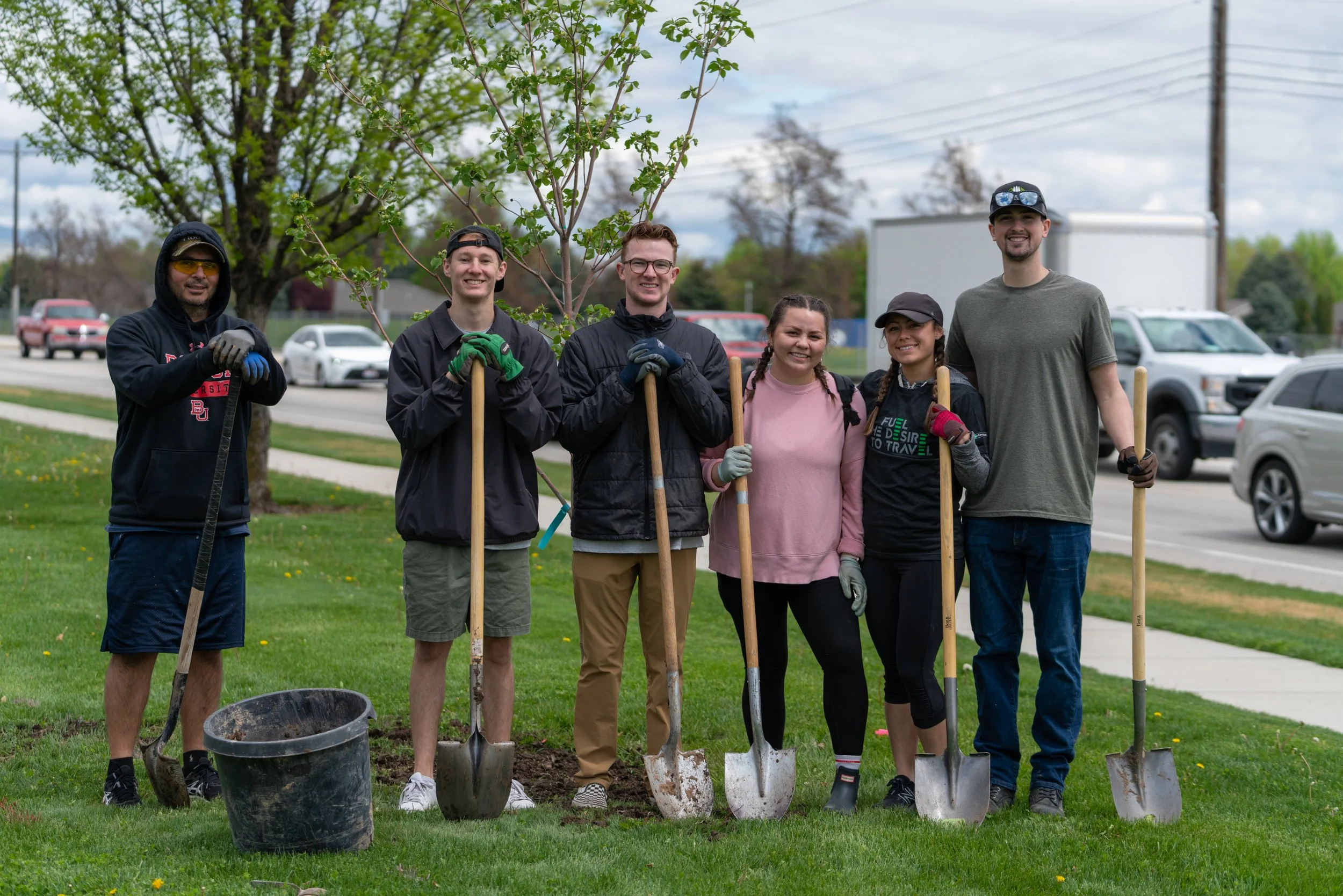 Group of six diverse young adults standing on grass with shovels, smiling, during a community gardening or tree planting event, with trees, cars, and a cloudy sky in the background.