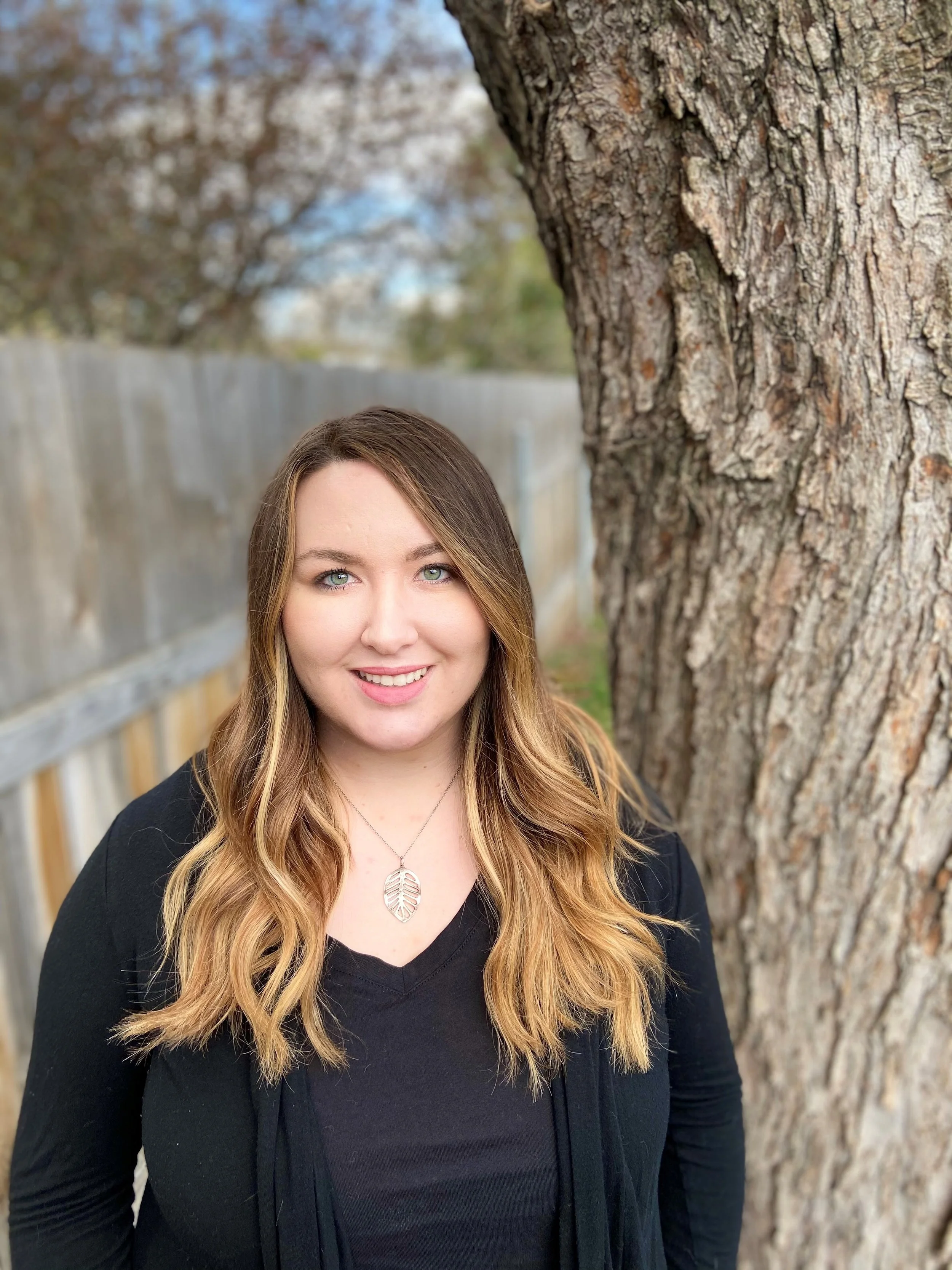 A young woman with long wavy blonde hair and blue eyes smiling outdoors near a tree and wooden fence.
