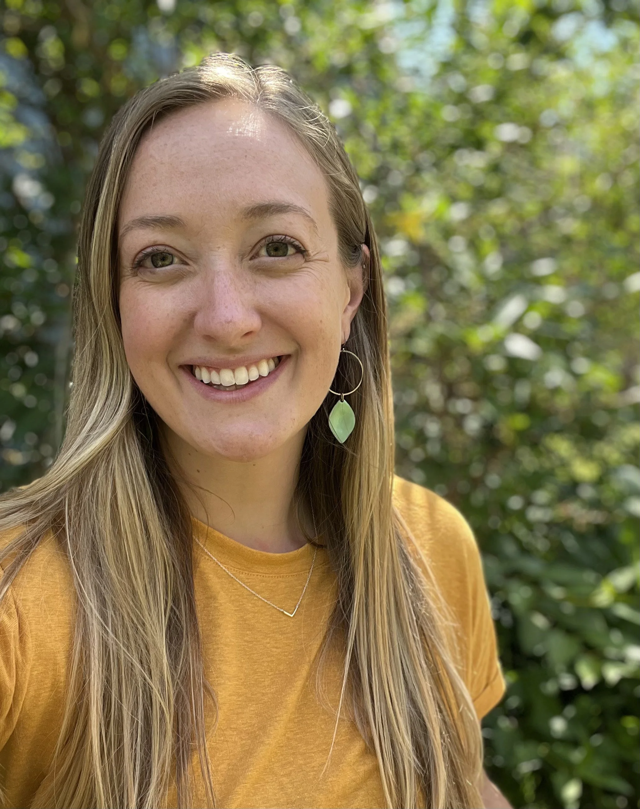 A woman smiling outdoors with trees in the background, wearing a yellow shirt and jewelry, including a necklace, hoop earrings, and a leaf-shaped earring.