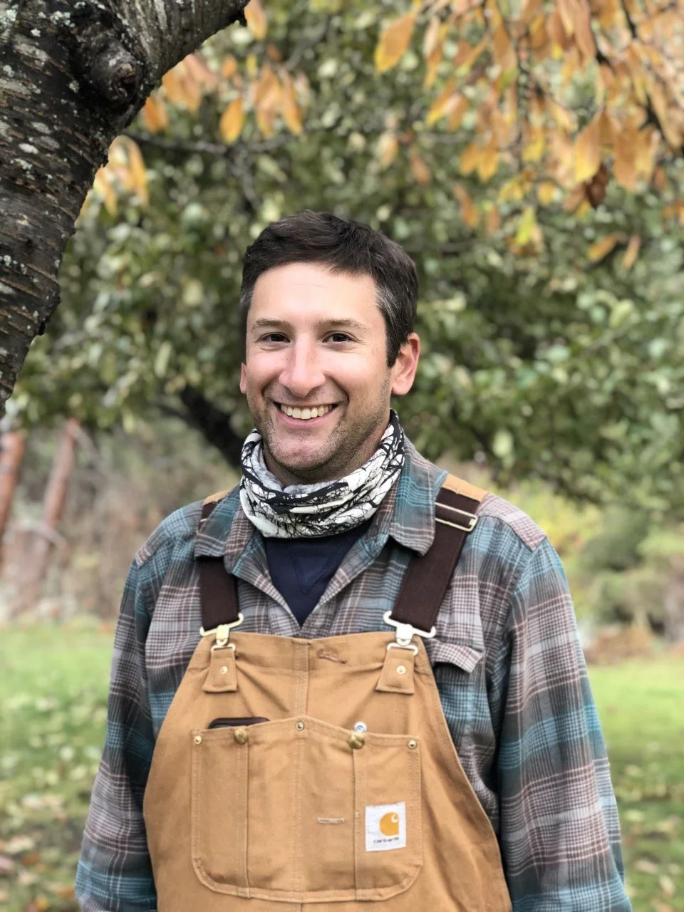 A smiling man outdoors in front of autumn-colored trees, wearing a plaid shirt, a Carhartt overalls, and a patterned scarf around his neck.