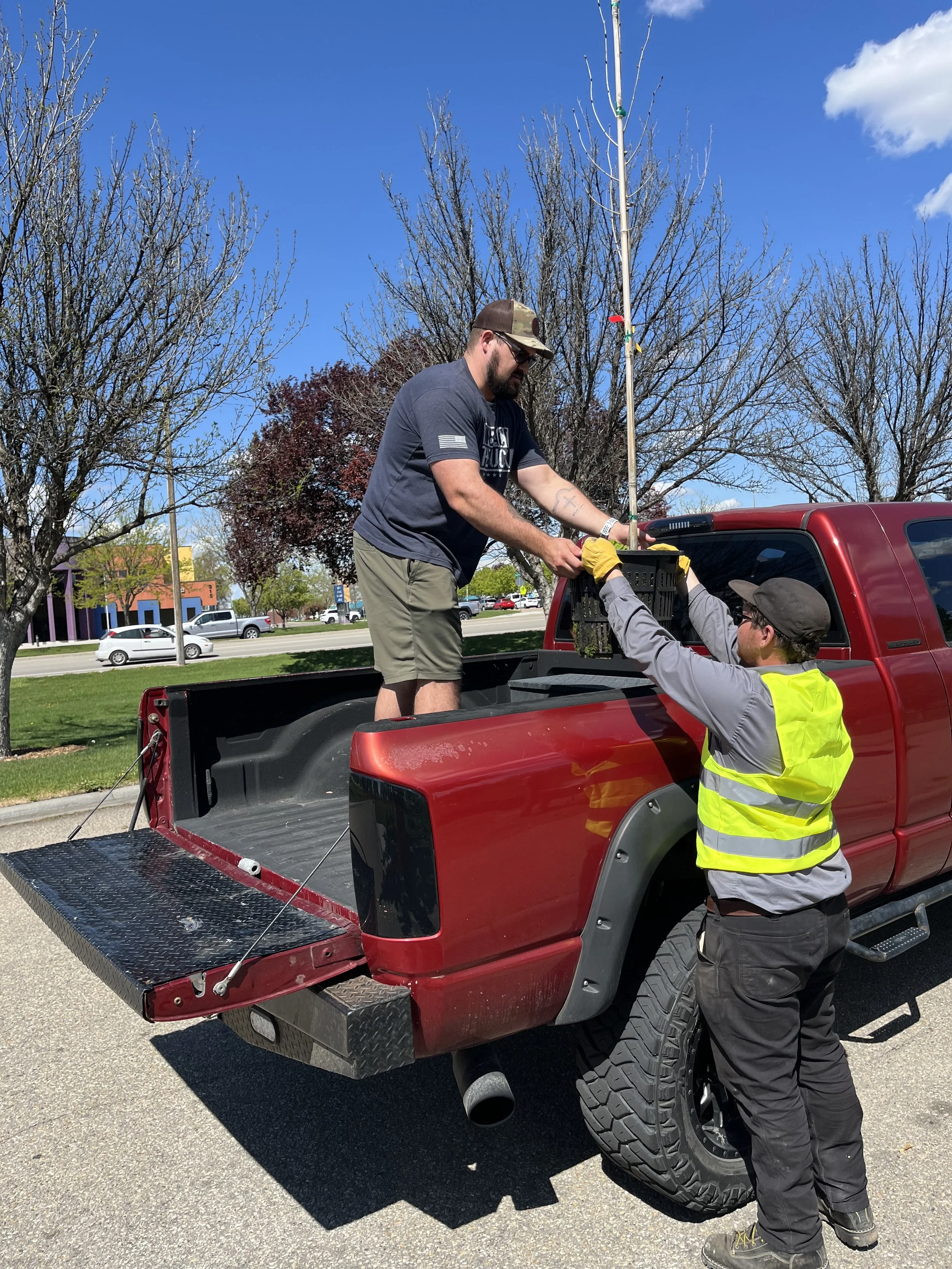 One man, standing on the truck bed, is wearing a gray t-shirt and camouflage cap, while the other, on the ground, is wearing a gray jacket and a yellow safety vest. They are outdoors on a sunny day with clear blue skies, leafless trees, and a parking lot with cars in the background.