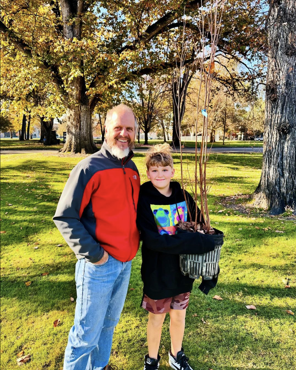 A man and young boy standing together in a park with fall trees in the background. The boy is holding a tree in a black pot.