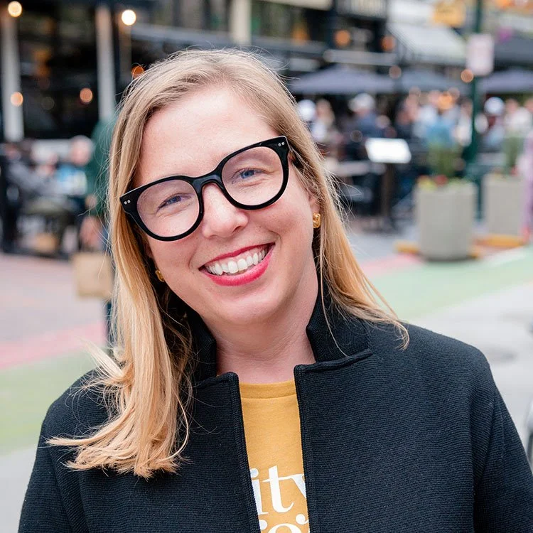 A smiling woman with blonde hair, large black glasses, wearing gold earrings, a yellow shirt, and a black jacket, standing outdoors in an urban setting.