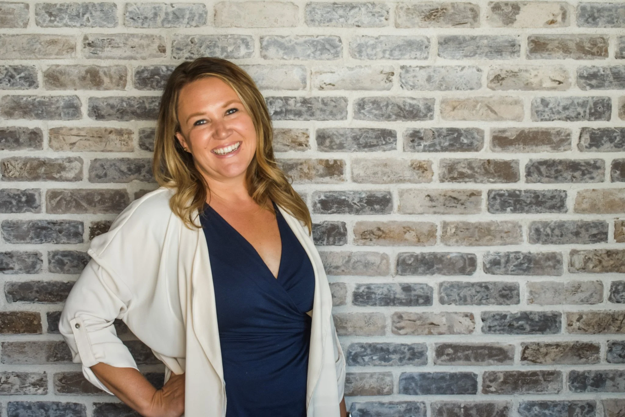 A woman with light brown, shoulder-length hair smiling, wearing a navy blue top and a white blazer, standing in front of a brick wall.