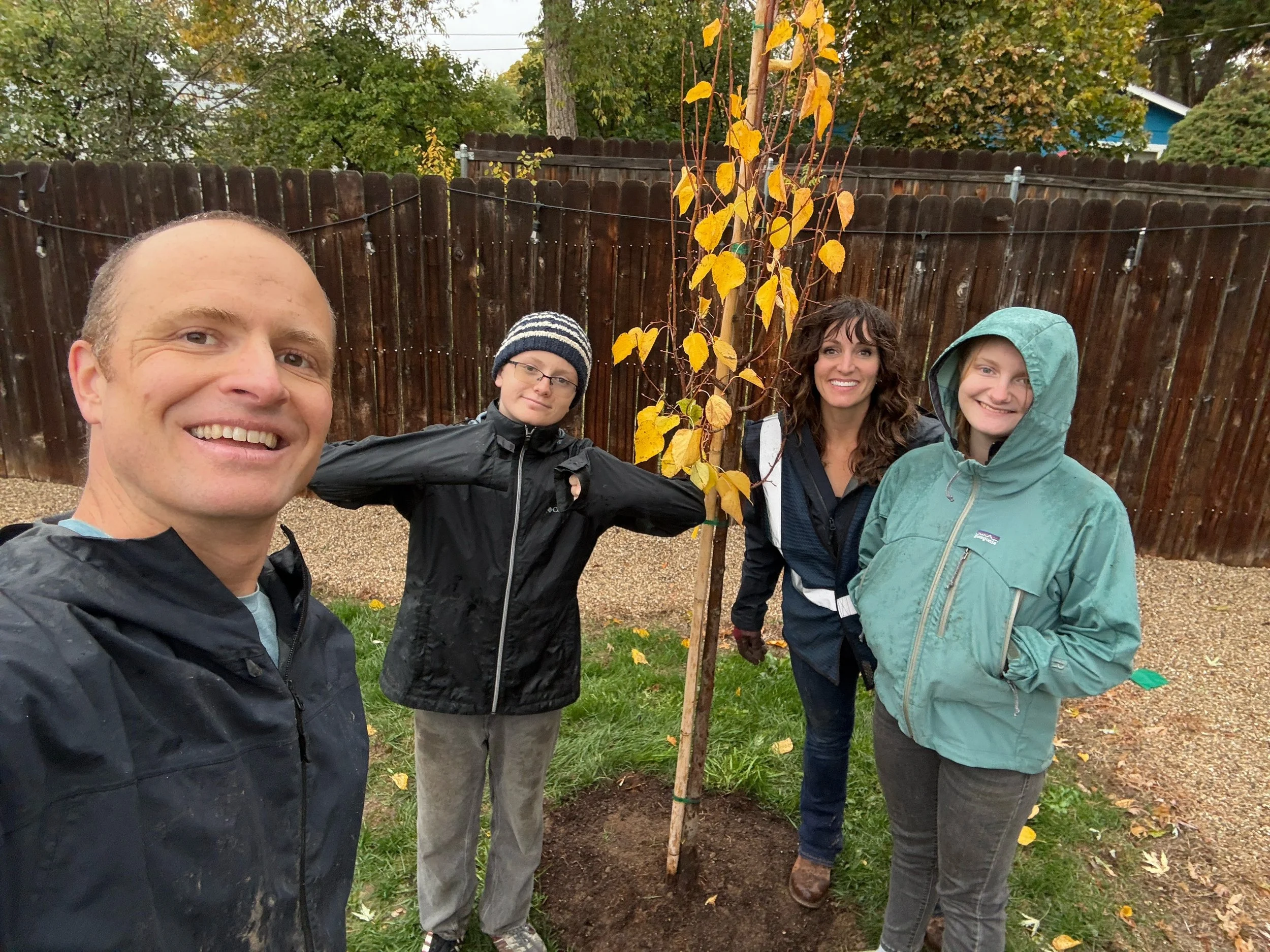 Four people standing outdoors in a backyard during fall, smiling and posing around a young tree with yellow leaves.
