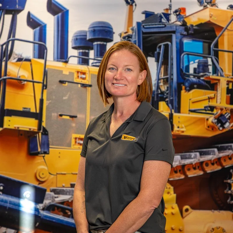 A woman with red hair standing in front of large yellow and blue construction equipment, smiling at the camera.