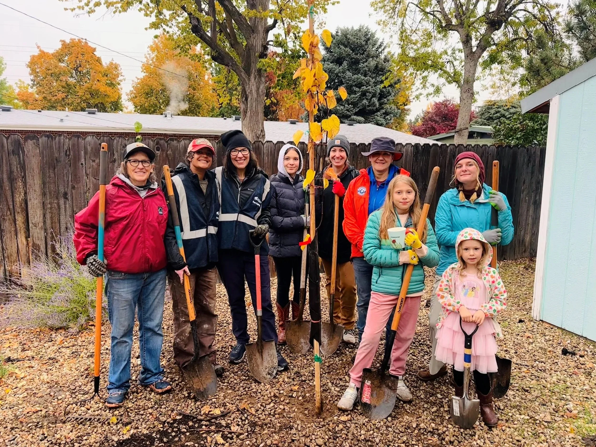 A group of ten people, including children, standing outdoors in a garden with a wooden fence and autumn trees in the background, holding shovels and working on planting a young tree.