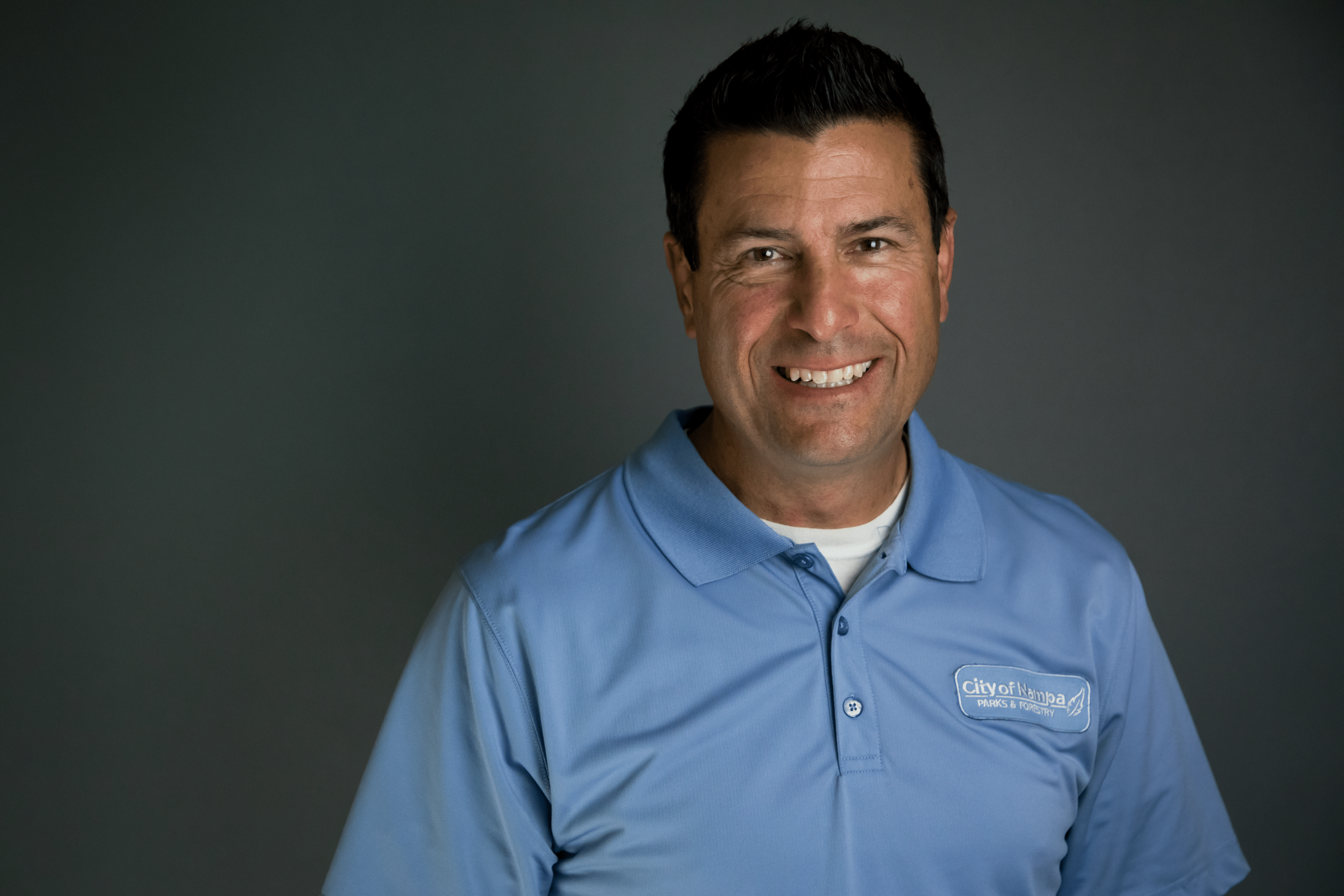 A man with dark hair smiling, wearing a blue polo shirt with a logo that says City of Napaa Parks & Forestry, against a dark background.