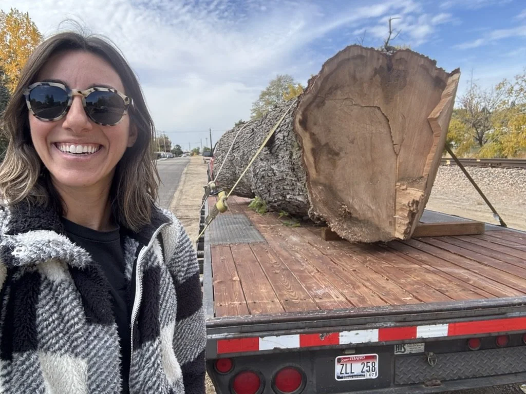 A woman with sunglasses smiling next to a large tree trunk on a flatbed truck.