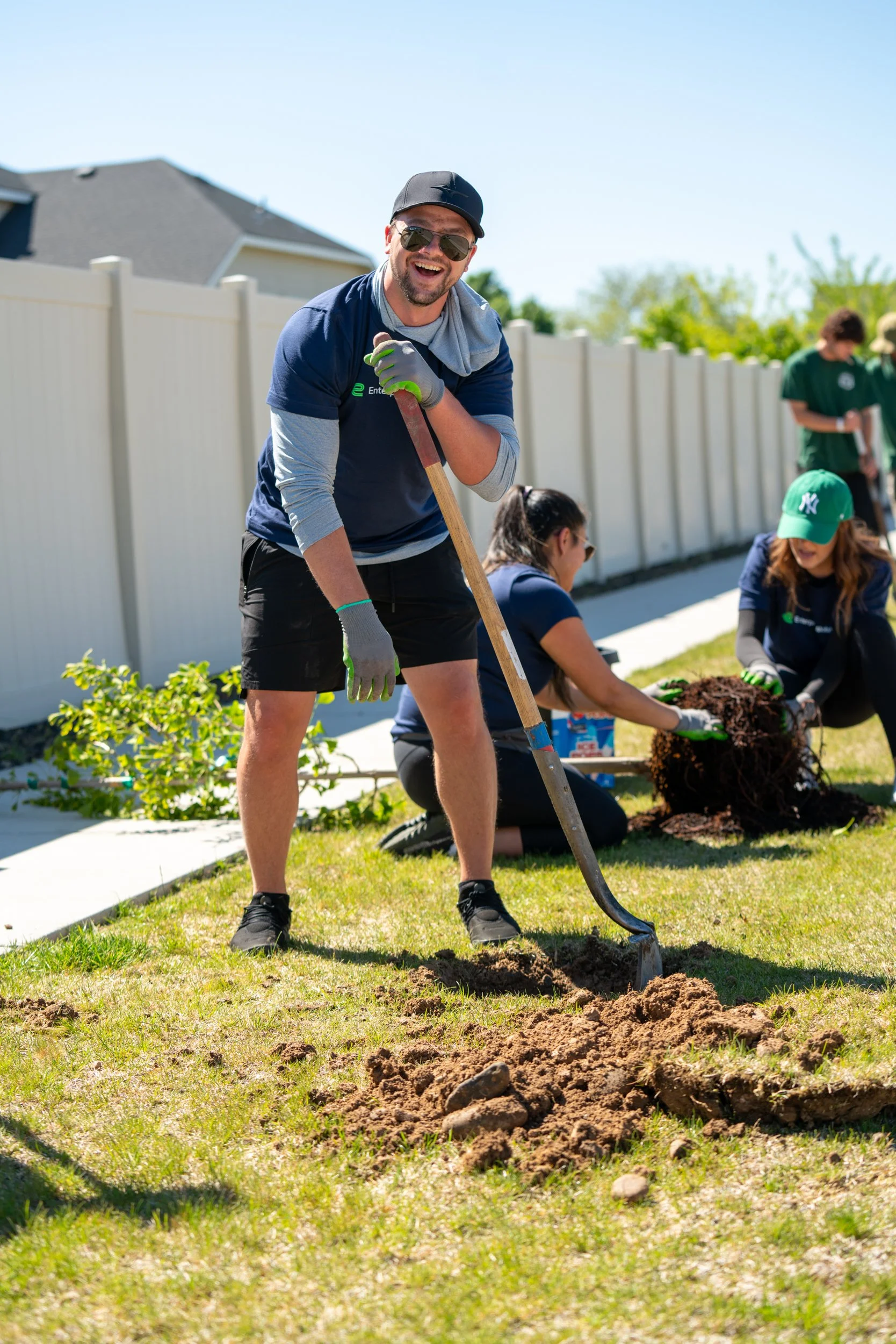Man smiling with sunglasses and gloves, digging a hole in a backyard with a shovel, surrounded by volunteers planting a tree.