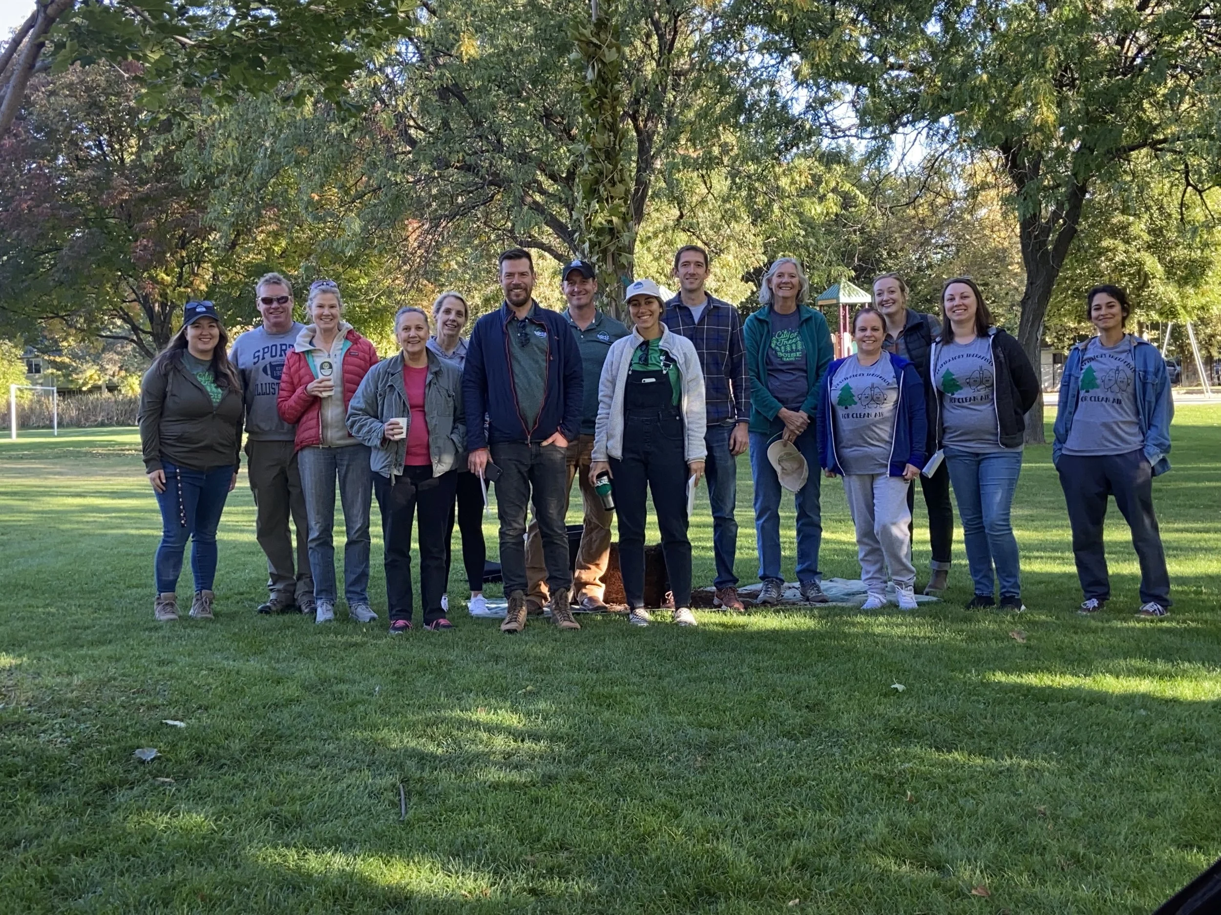 Group of people standing outdoors on a grassy field, posing for a photo near a large tree, with some wearing matching gray t-shirts with a logo, in a park setting.