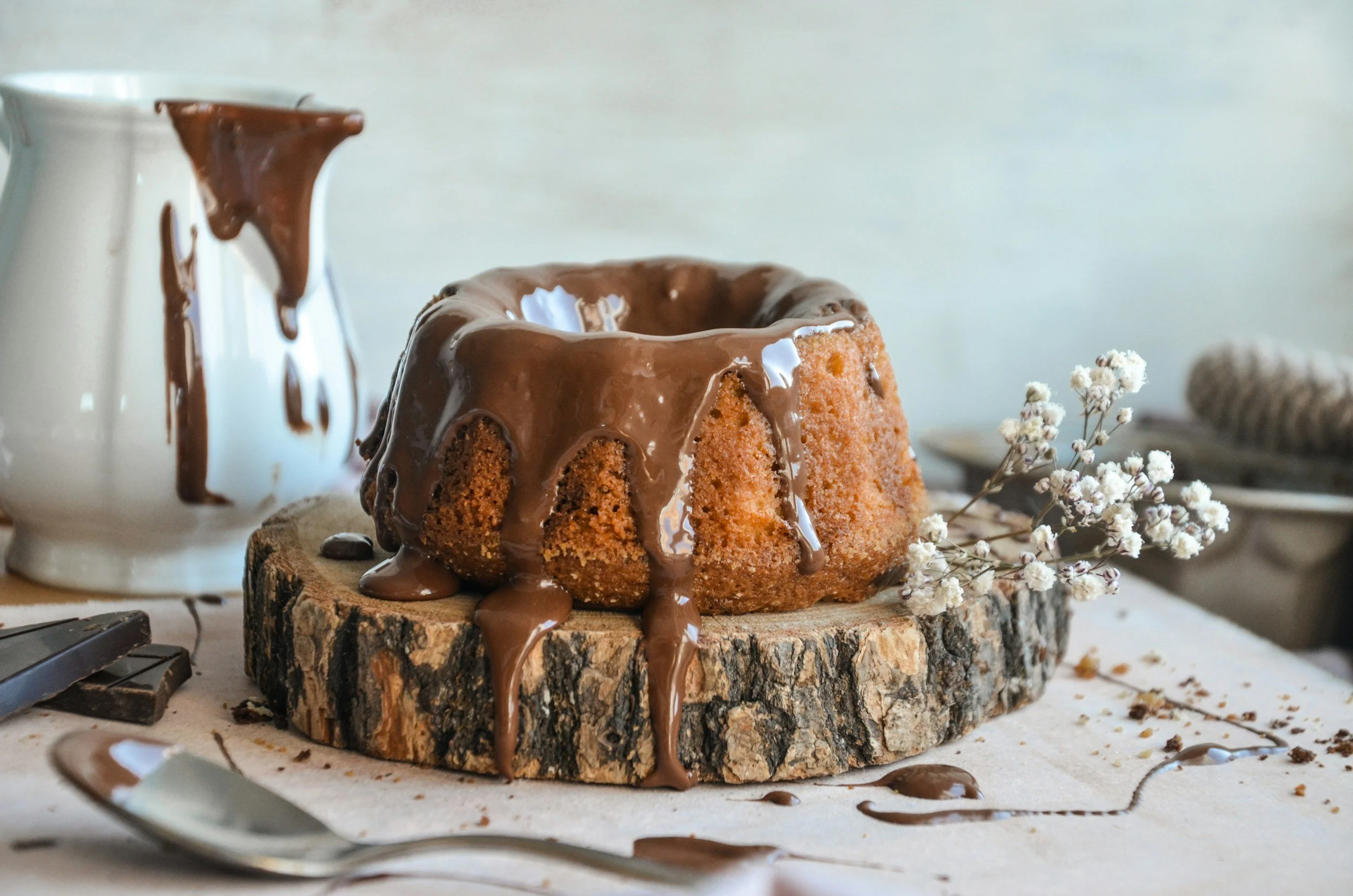 Chocolate-glazed bundt cake on a wooden slab with white flower decoration, surrounding utensils, and a ceramic pitcher with chocolate drips in the background.