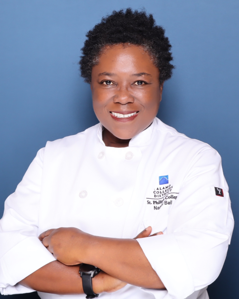 A smiling woman in a white chef's coat with a logo, standing with her arms crossed against a blue background.