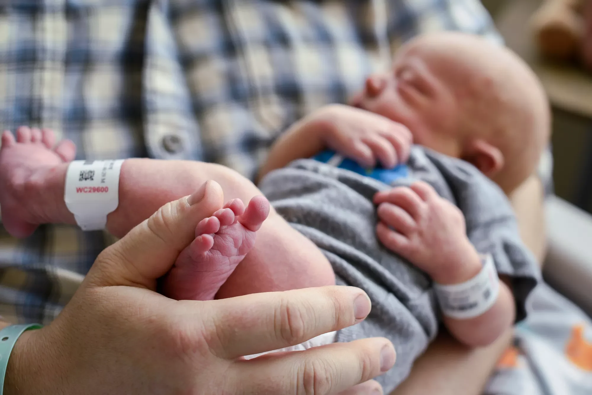A newborn baby with tiny feet is being held by a person wearing a plaid shirt.