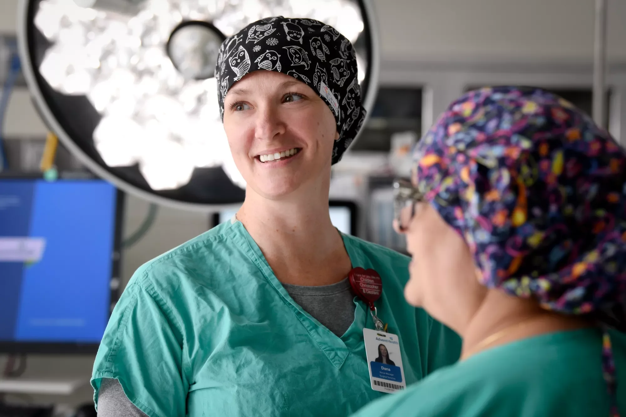 Two smiling nurses in scrubs and head coverings converse in a hospital room.