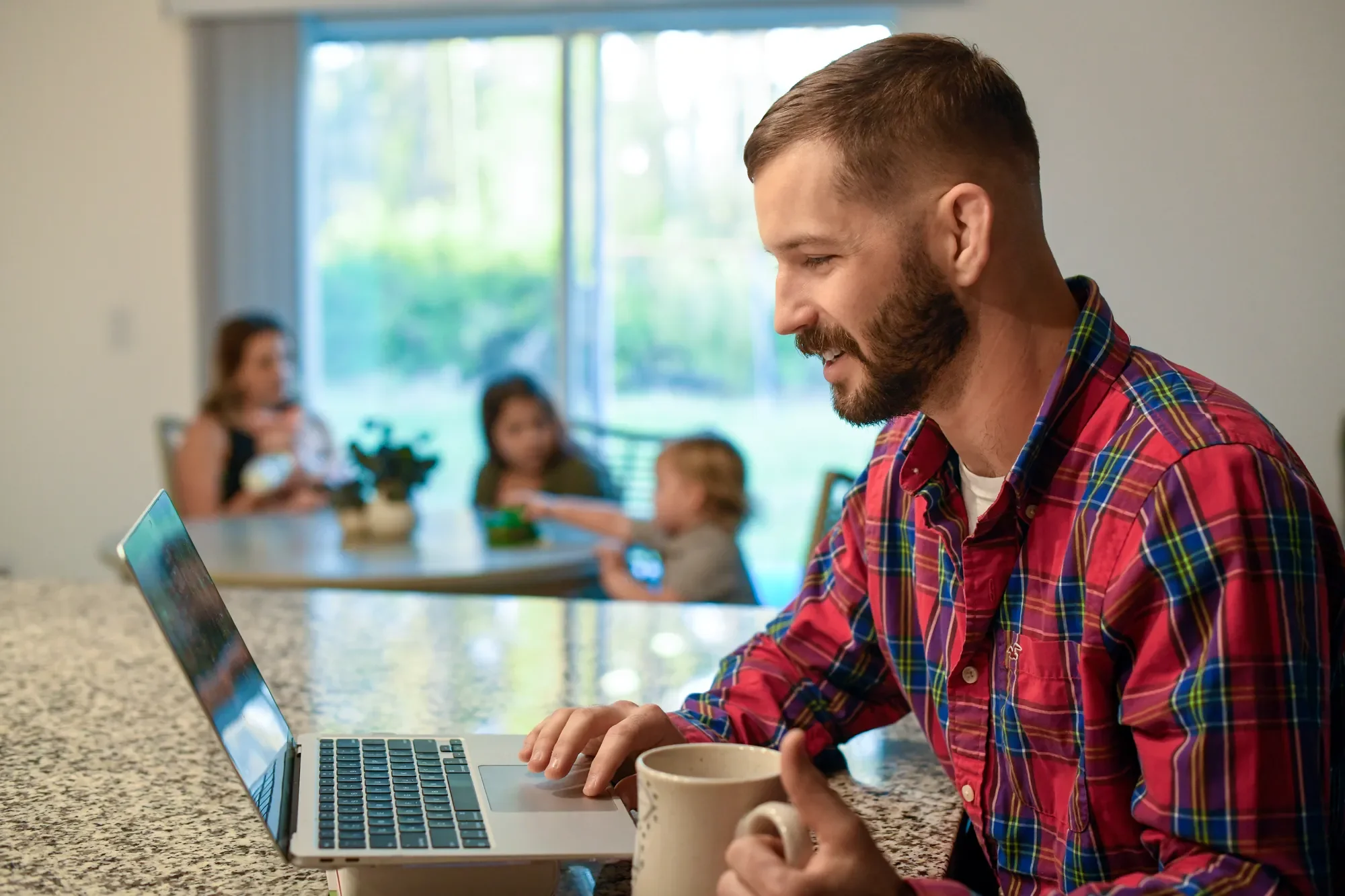 Man in plaid shirt working on laptop at home with family in background.