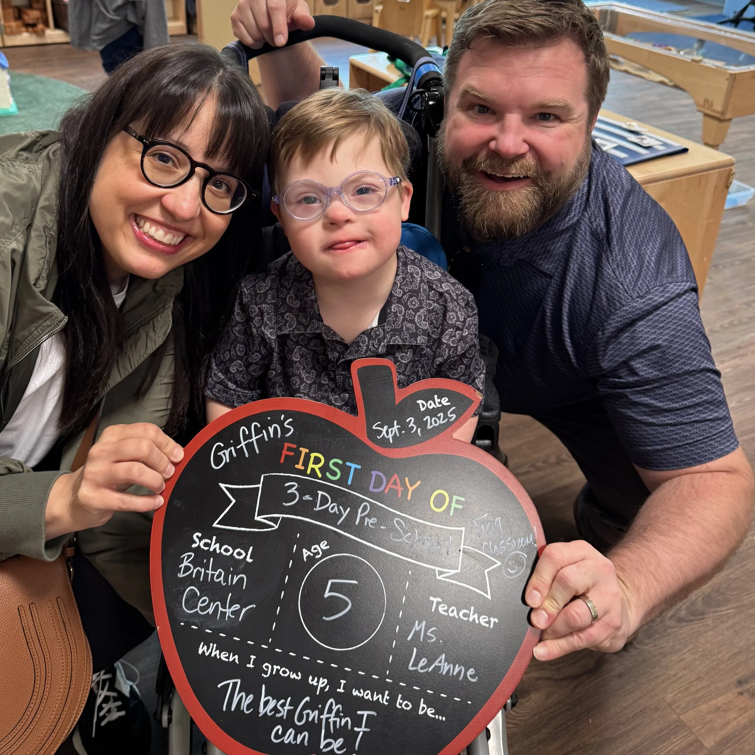 Griffin smiles with his parents while holding a sign celebrating his first day of preschool at Britain Development.