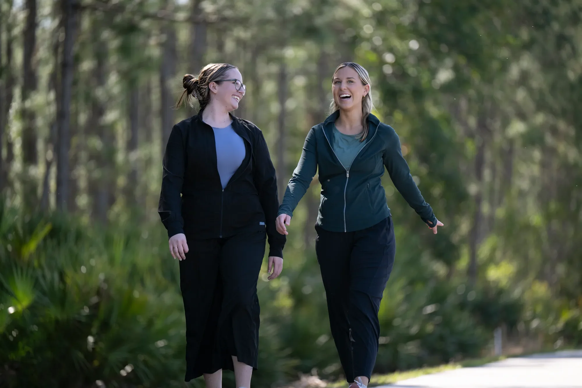 Two women walking and laughing on a path in a wooded area, promoting a sense of community and joy.