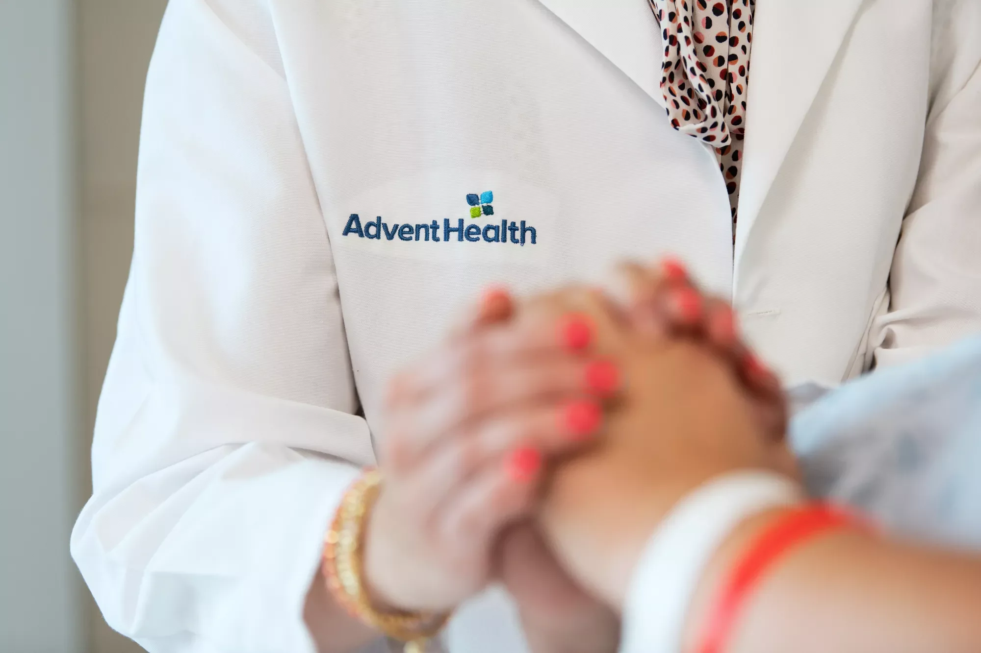 A close-up of a doctor and patient holding hands, with the AdventHealth logo visible on the doctor's coat.