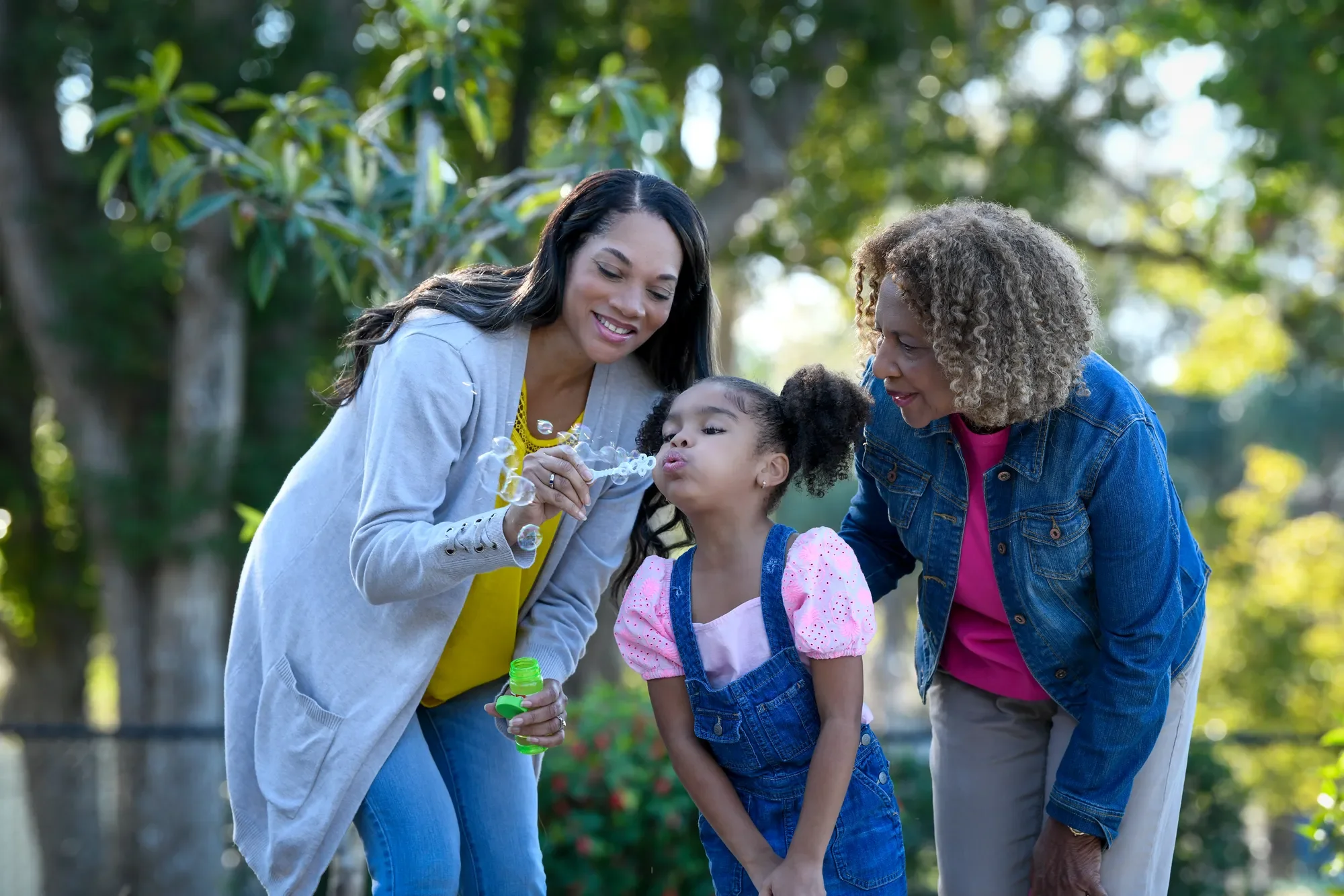 Three generations of women smiling and blowing bubbles in a park.