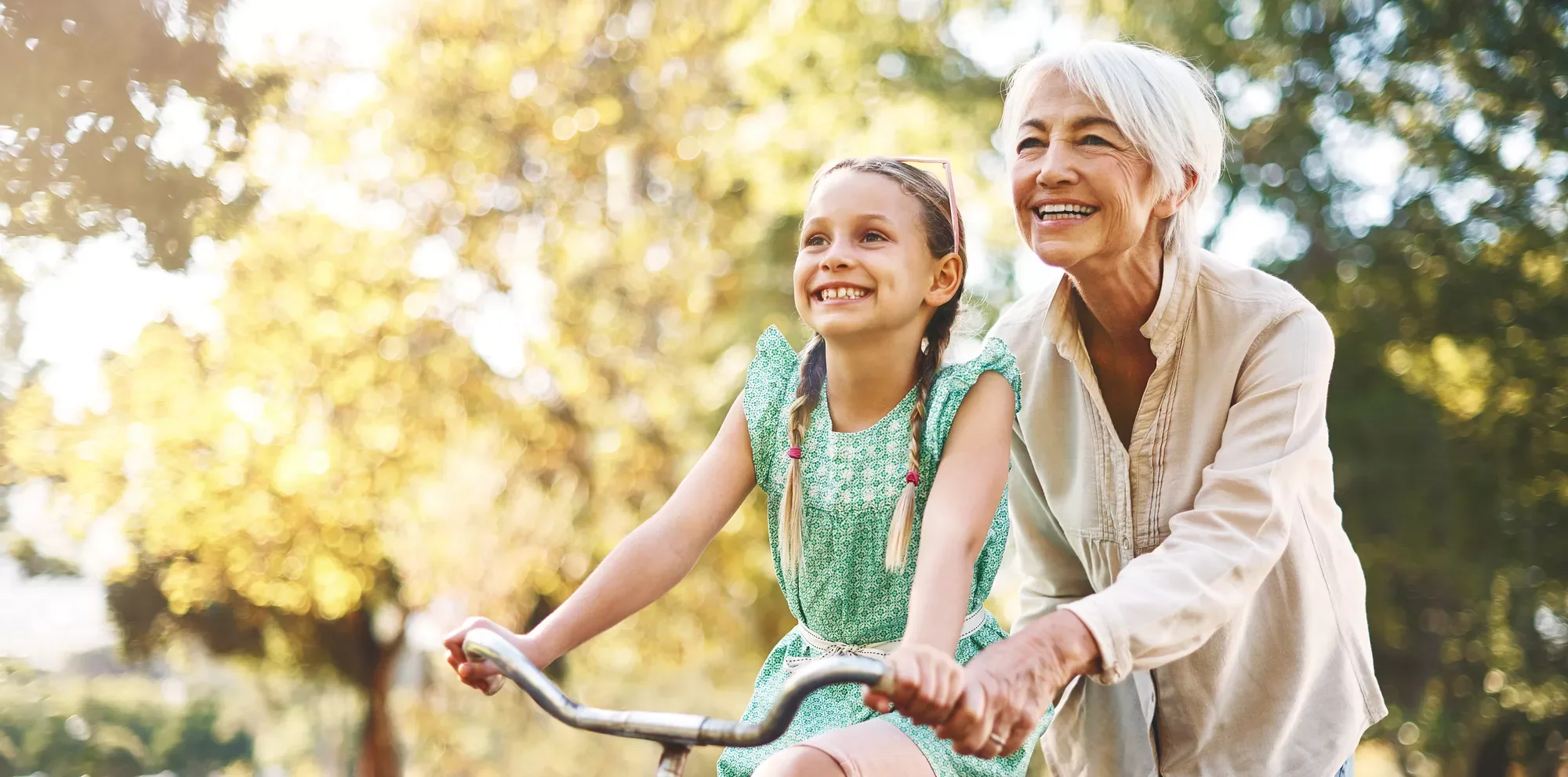 An older woman supports a smiling young girl as she learns to ride a bike outdoors, capturing a moment of care and connection.