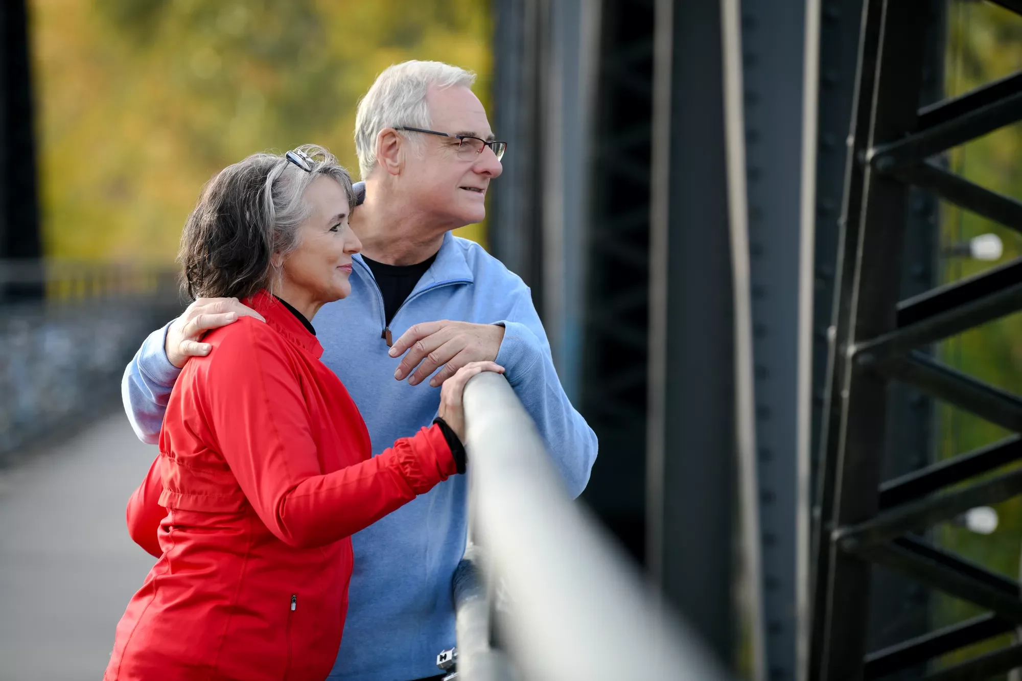 An older couple stands together on a bridge, sharing a quiet moment of connection, support, and well-being.