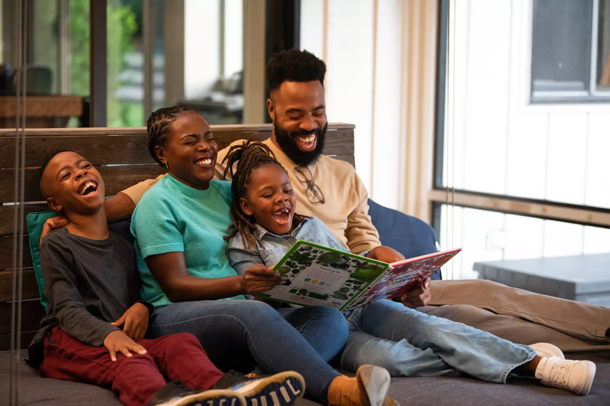 A family of four, consisting of two adults and two children, are sitting together on a couch, laughing and reading a book.