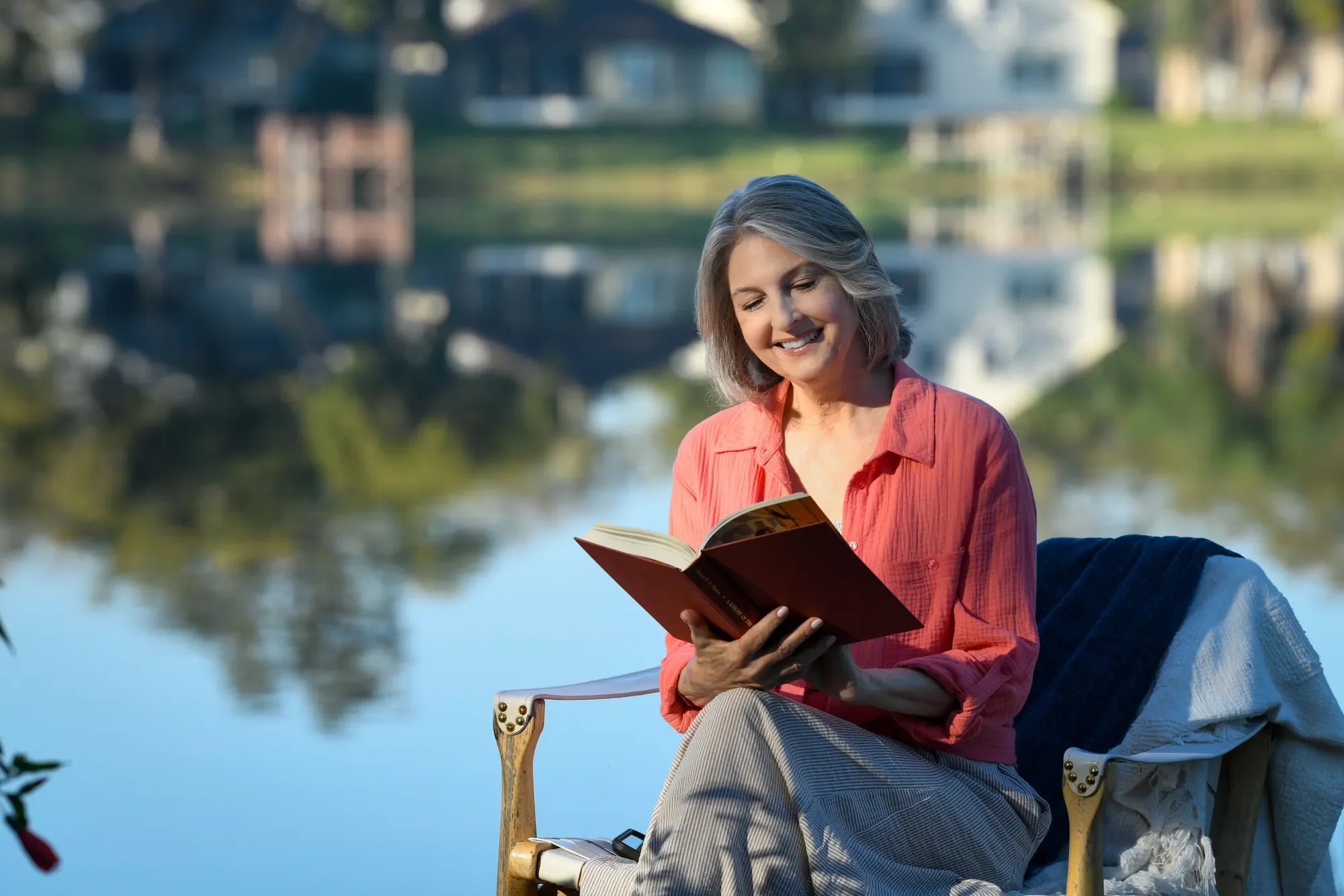 A woman sits by a calm lakeside reading a book, reflecting a moment of mental well-being, peace, and emotional balance.