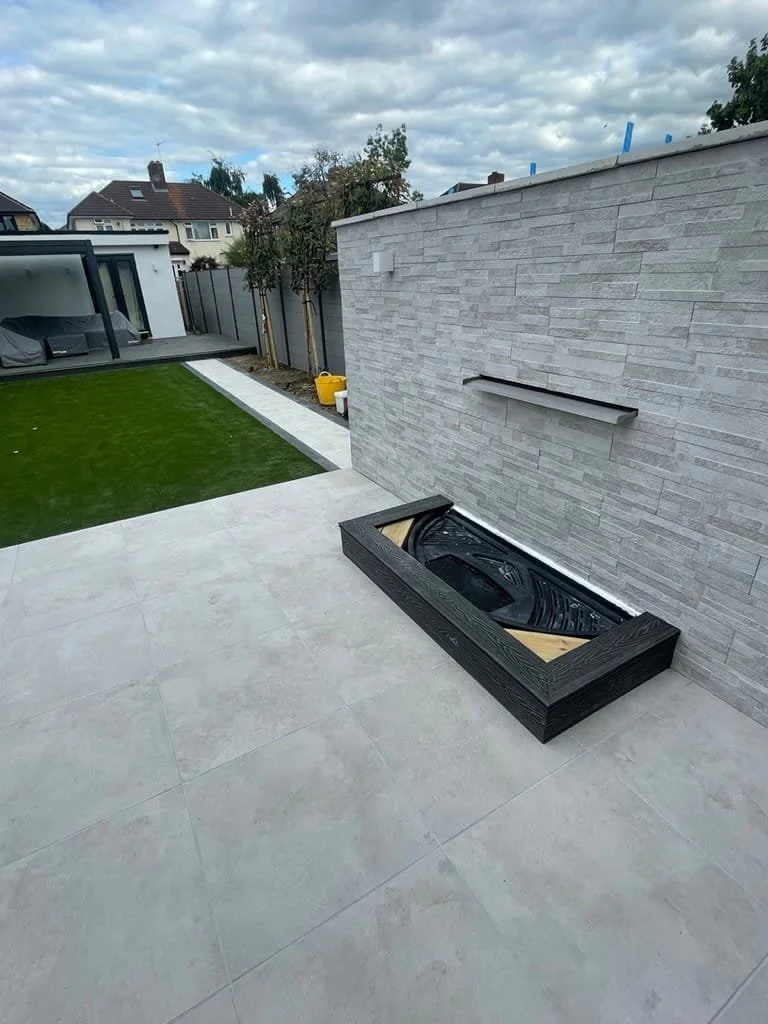 Newly finished backyard with concrete patio, artificial grass, and a modern gray stone wall. A built-in hot tub is installed in a wooden frame on the patio. The yard is surrounded by a fence, with houses and trees in the background under a cloudy sky.