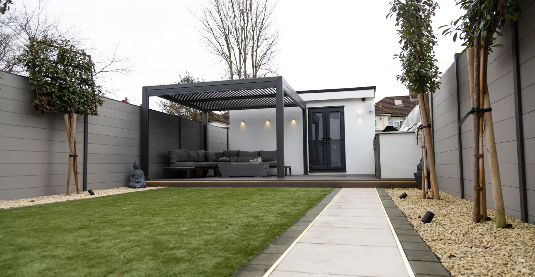 Modern backyard with a gray privacy fence, a grassy lawn, and a pathway leading to a deck with seating and a pergola.