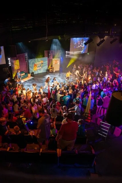 Large indoor event with a stage and audience, colorful lighting, and people dancing and watching entertainment.