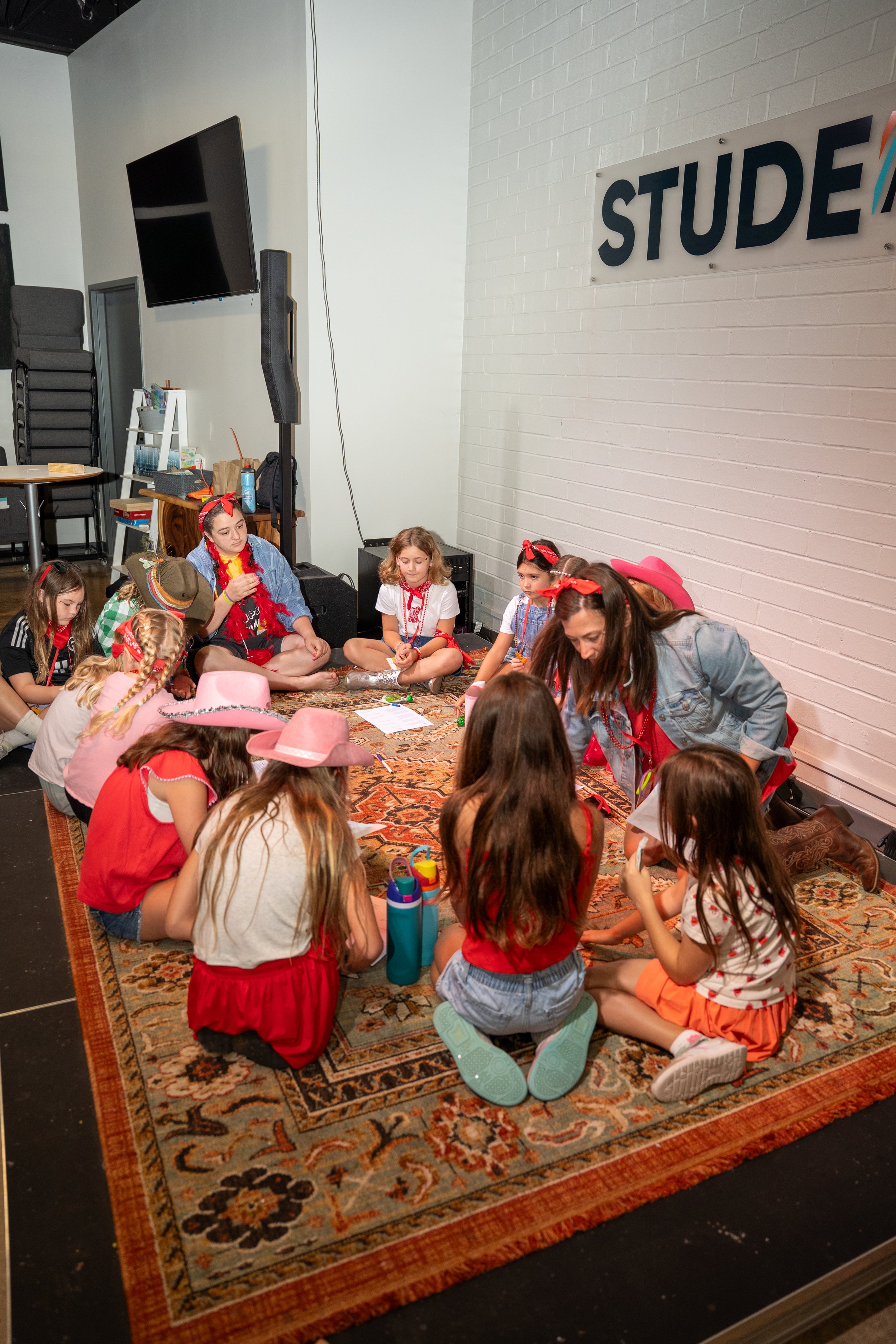 A group of young girls, dressed in casual and Western-themed clothing, sitting on a decorative rug in a circle while participating in a group activity in a room with a white brick wall and a sign that reads 'STUDE'.