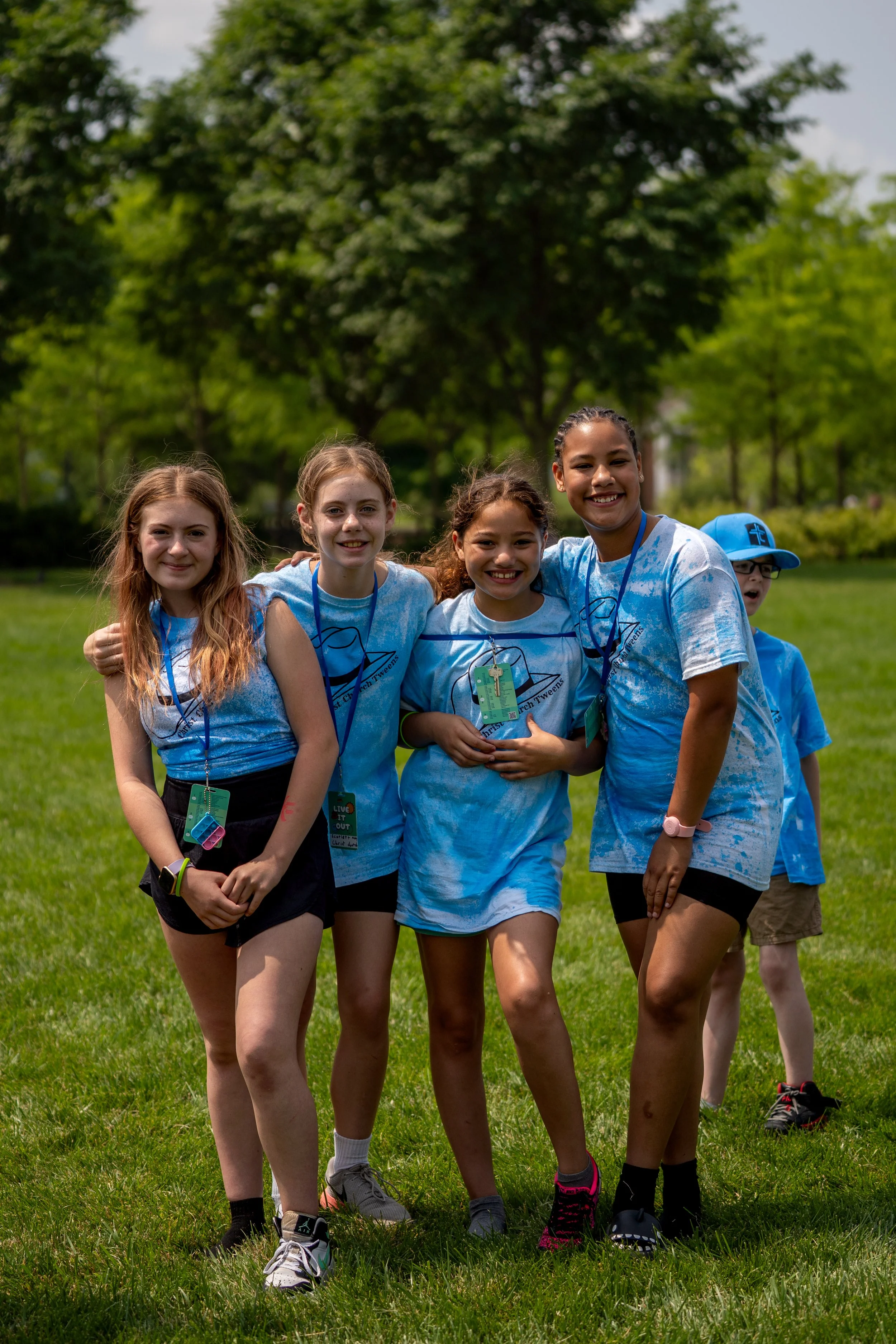 Four young girls standing together outdoors on a grassy field, smiling, with trees in the background.