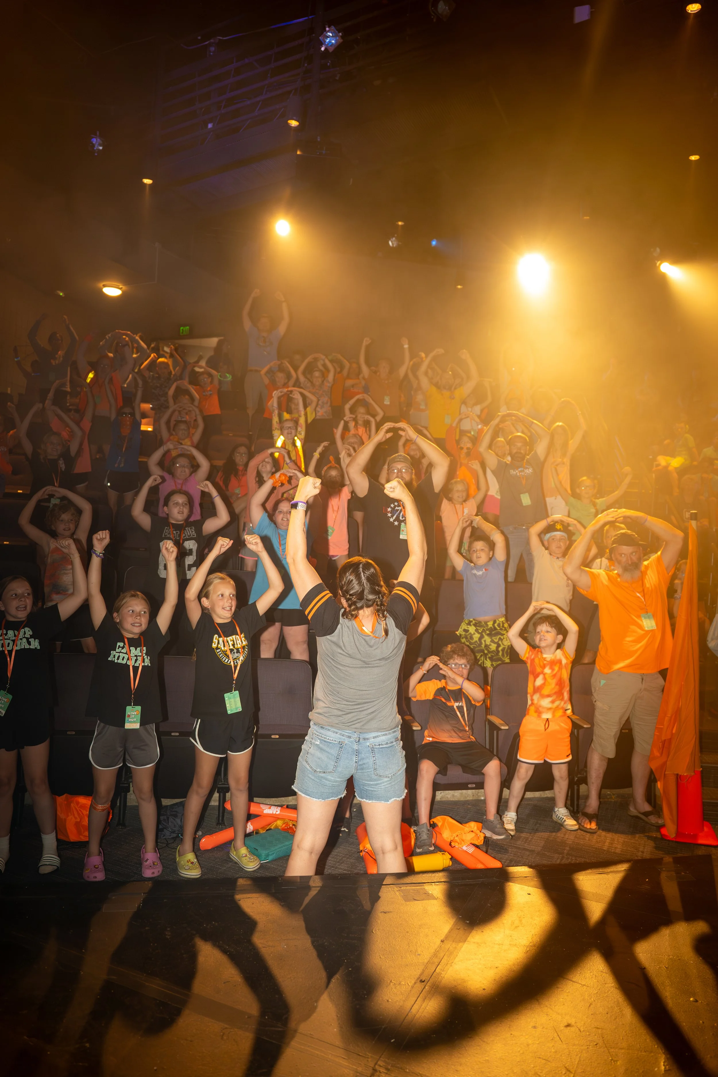 A group of children and adults on stage participating in a group activity, with many raising their arms and making heart shapes with their hands, illuminated by stage lighting.