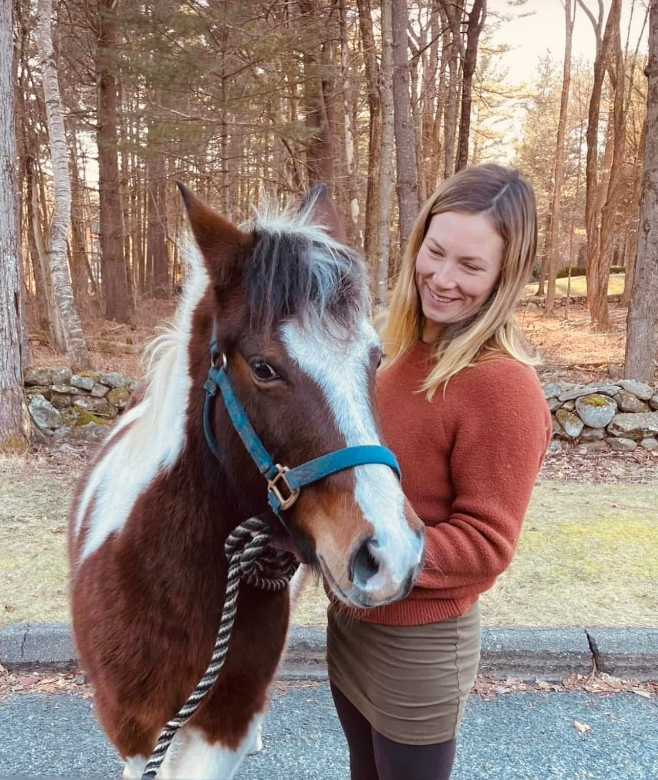 A woman smiling while holding a brown and white horse on a leash outdoors in a wooded area during sunset.