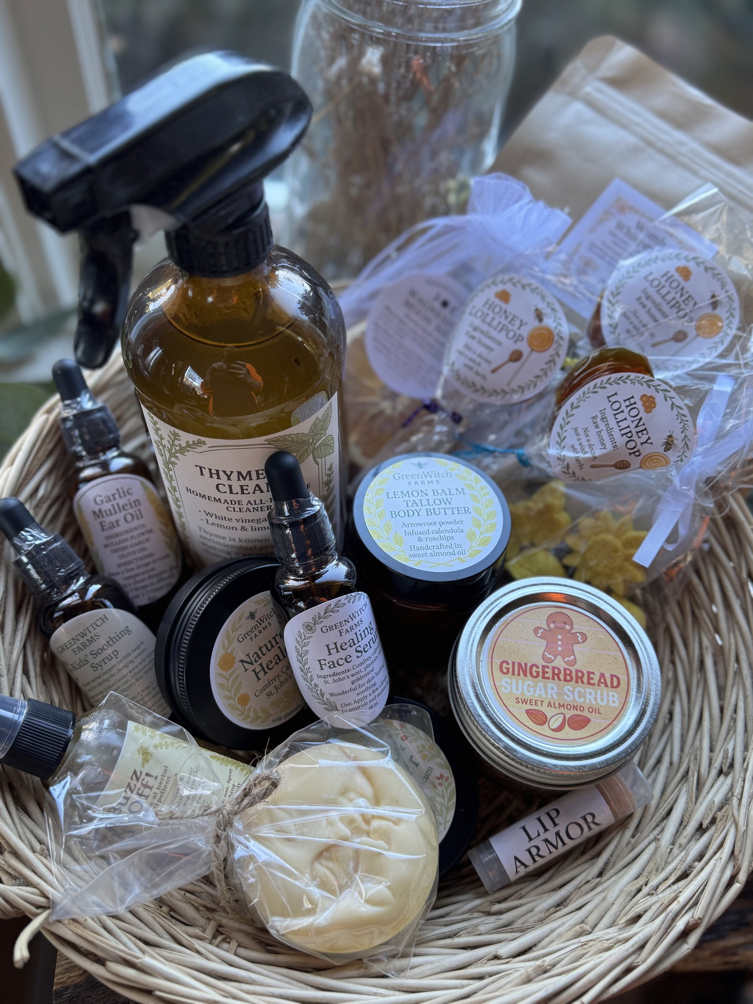 A wicker basket filled with natural and homemade skincare products, including honey lotion, gingerbread sugar scrub, lip balm, healing face serum, kid soothing syrup, garlic Mullein ear oil, and lemon balm body butter, with some items wrapped in clear plastic or glass bottles.
