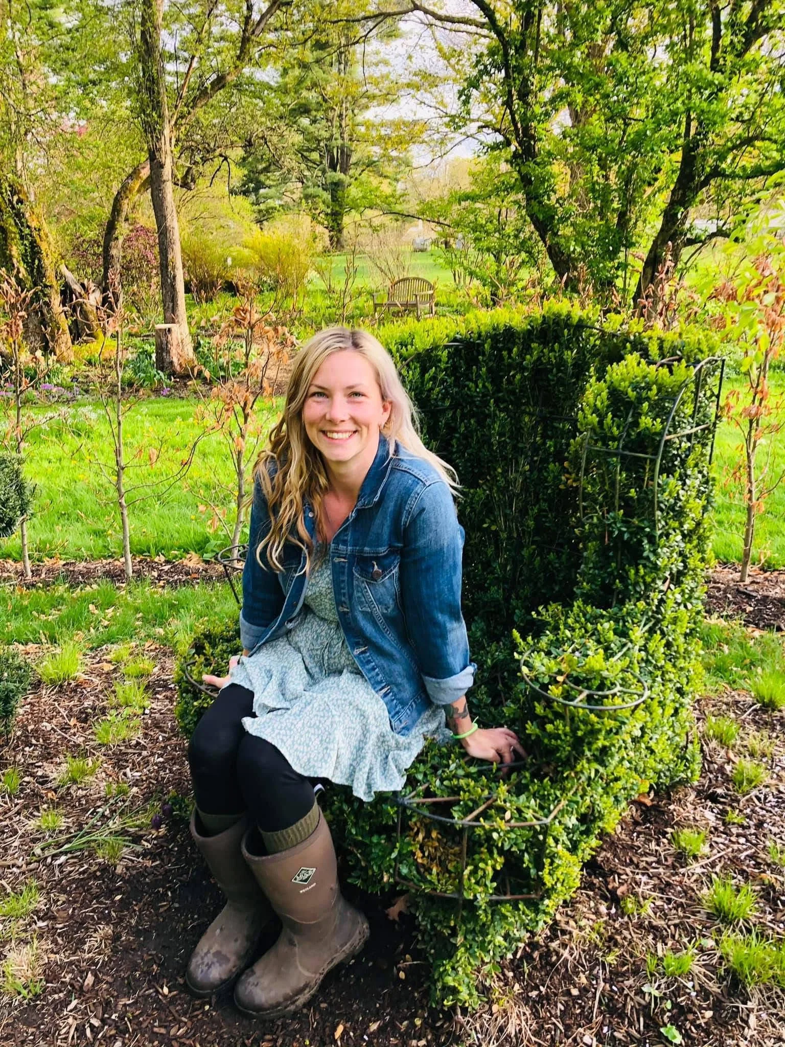 A woman sitting on a small, shaped hedge in a lush, green garden with trees and a bench in the background, smiling at the camera.