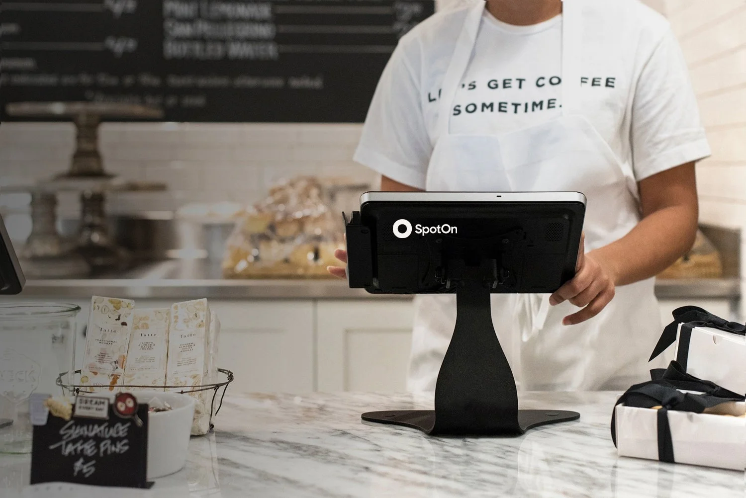A person standing behind a counter with a black Point of Sale device labeled 'SpotOn'. The person is wearing a white t-shirt with partially visible text and an apron. On the counter, there are small packets, a small chalkboard sign advertising 'Sausage Take Aways $5', and some wrapped items. In the background, a countertop with baked goods and a blackboard menu is visible.