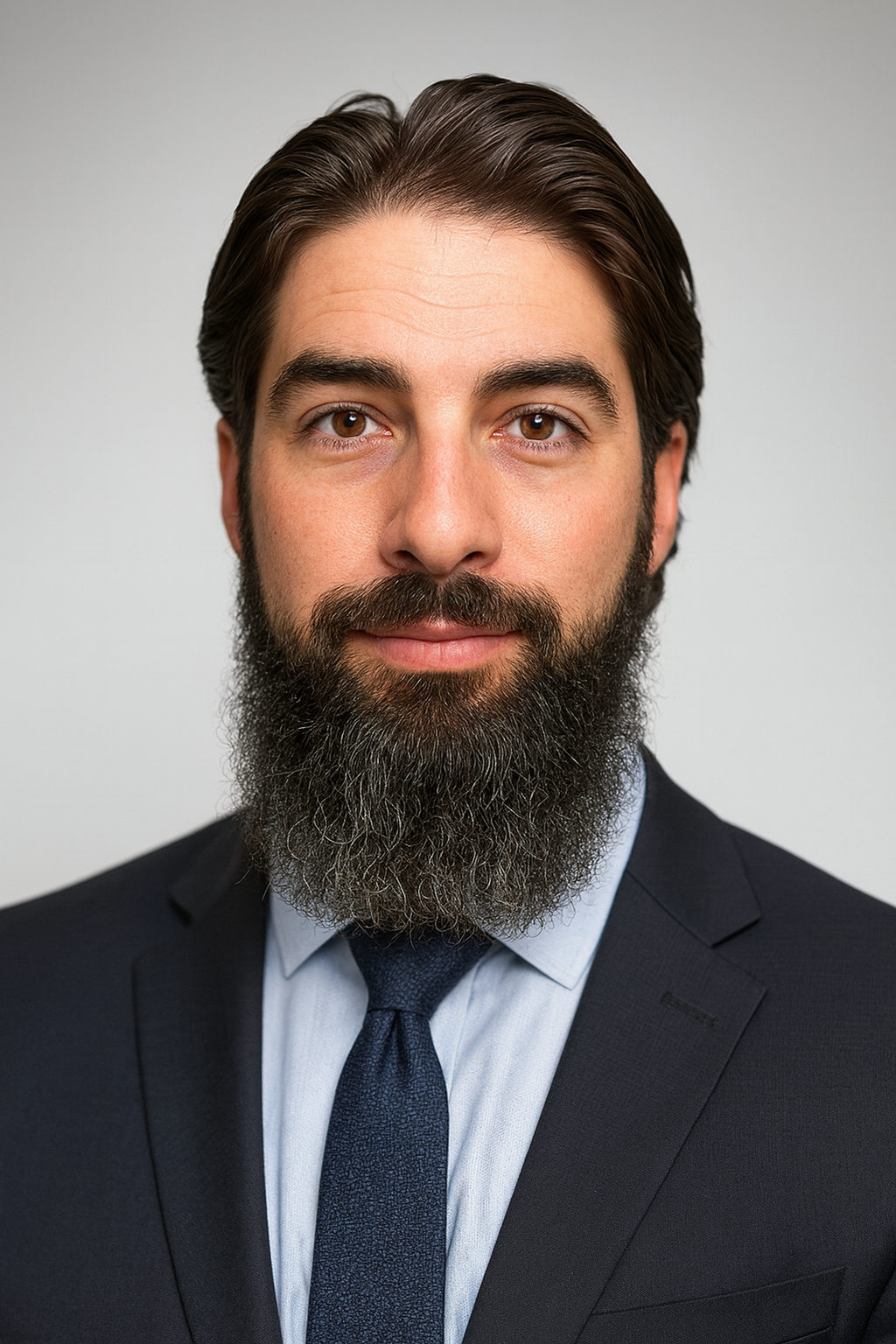 Headshot of a man with brown hair, brown eyes, and a full beard, wearing a dark suit, light shirt, and dark tie, against a gray background.
