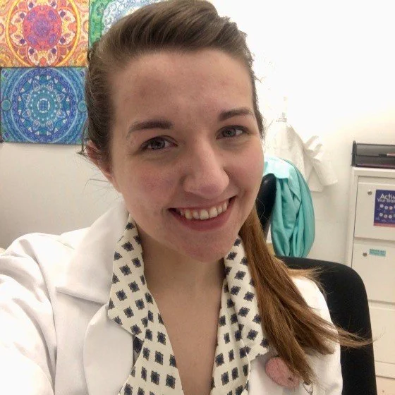 A young woman with light brown hair tied in a ponytail, wearing a white coat over a patterned shirt, smiling in an office or clinic setting with colorful artwork on the wall behind her.