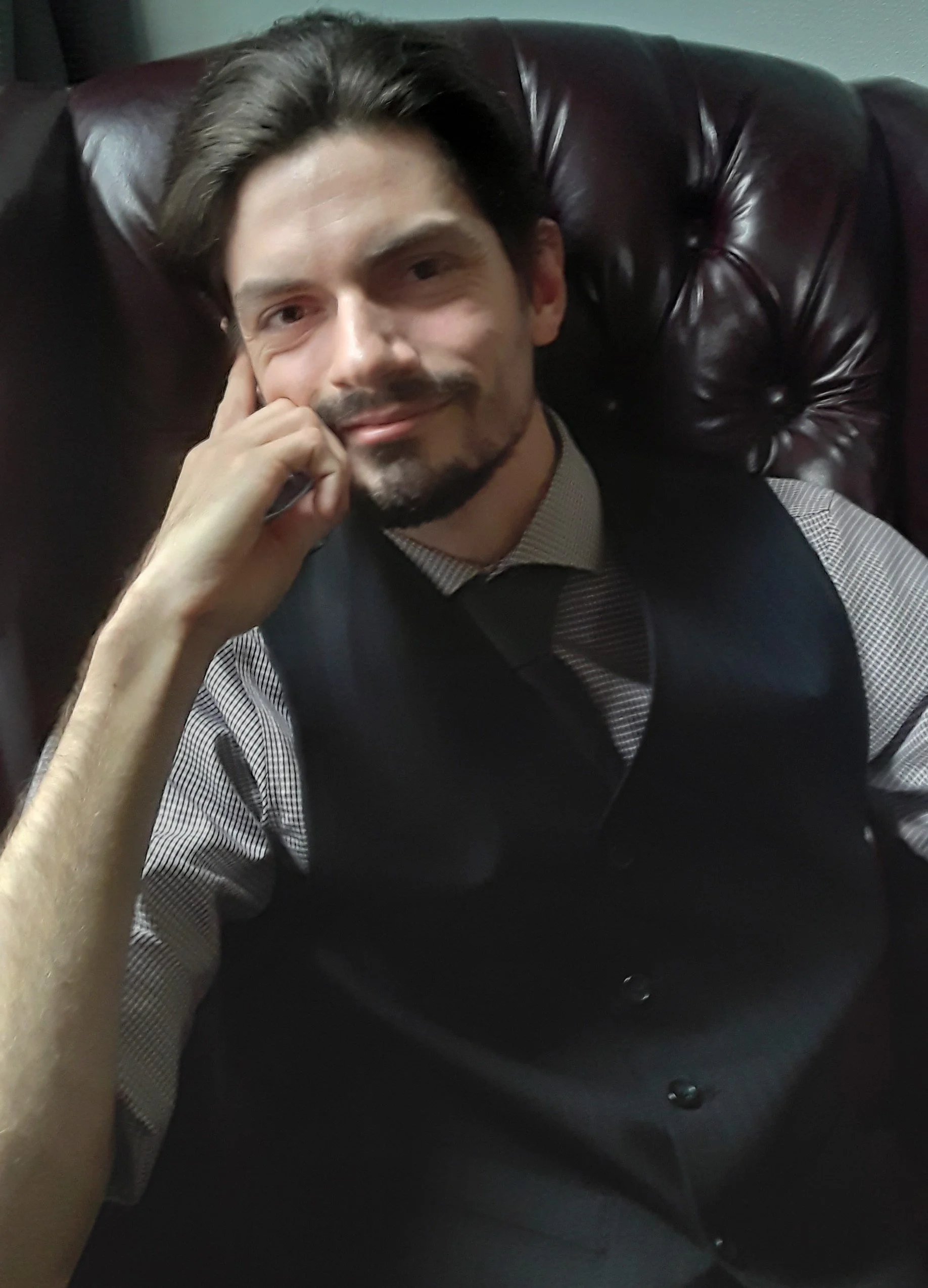 A man with dark hair and facial hair sitting on a brown leather chair, wearing a gray and white checked shirt and a black vest, smiling and resting his head on his hand.