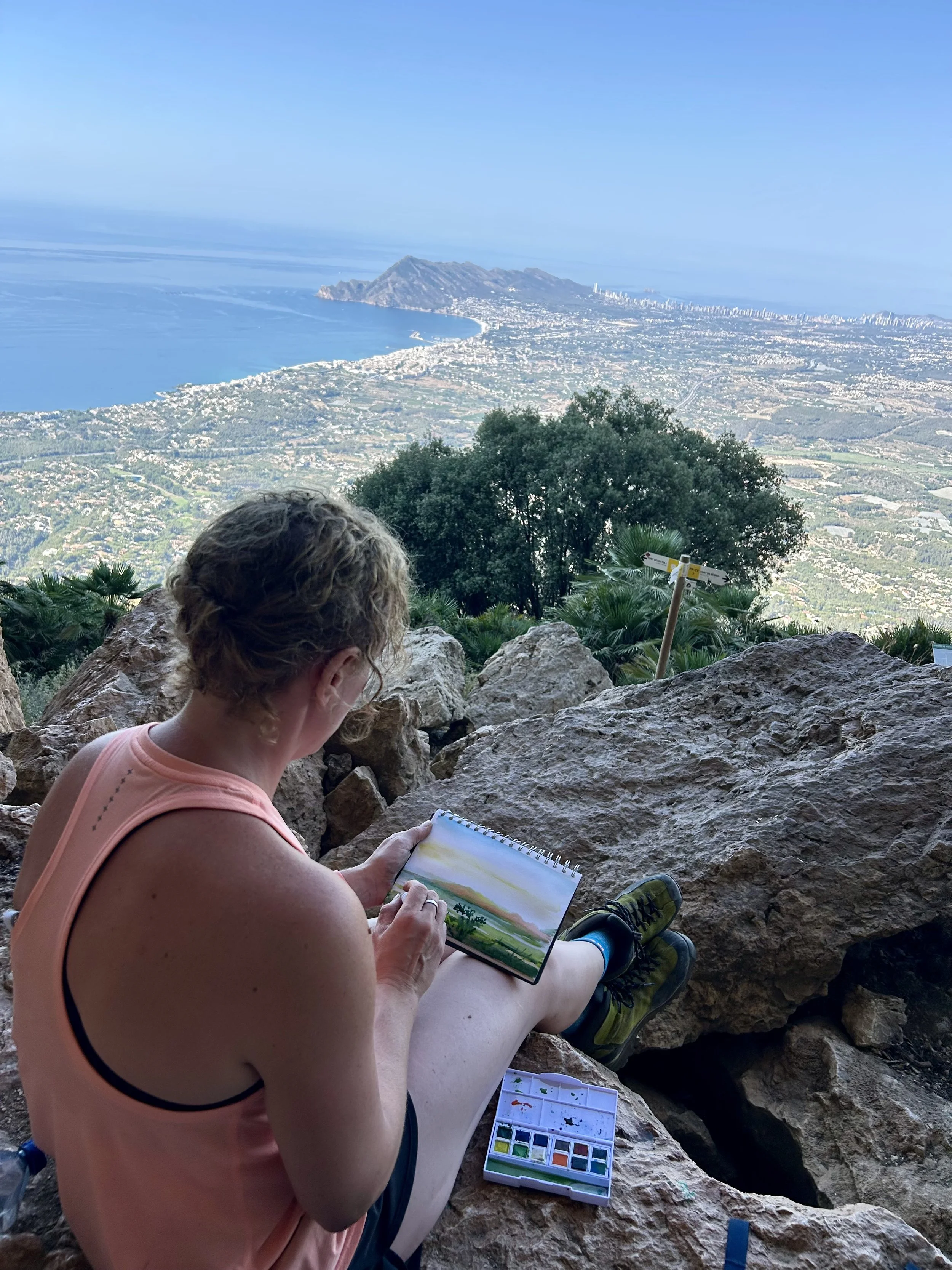Cat Goward painting a landscape with watercolour paints while sitting on rocks with a scenic view of mountains, city, and ocean in the background.