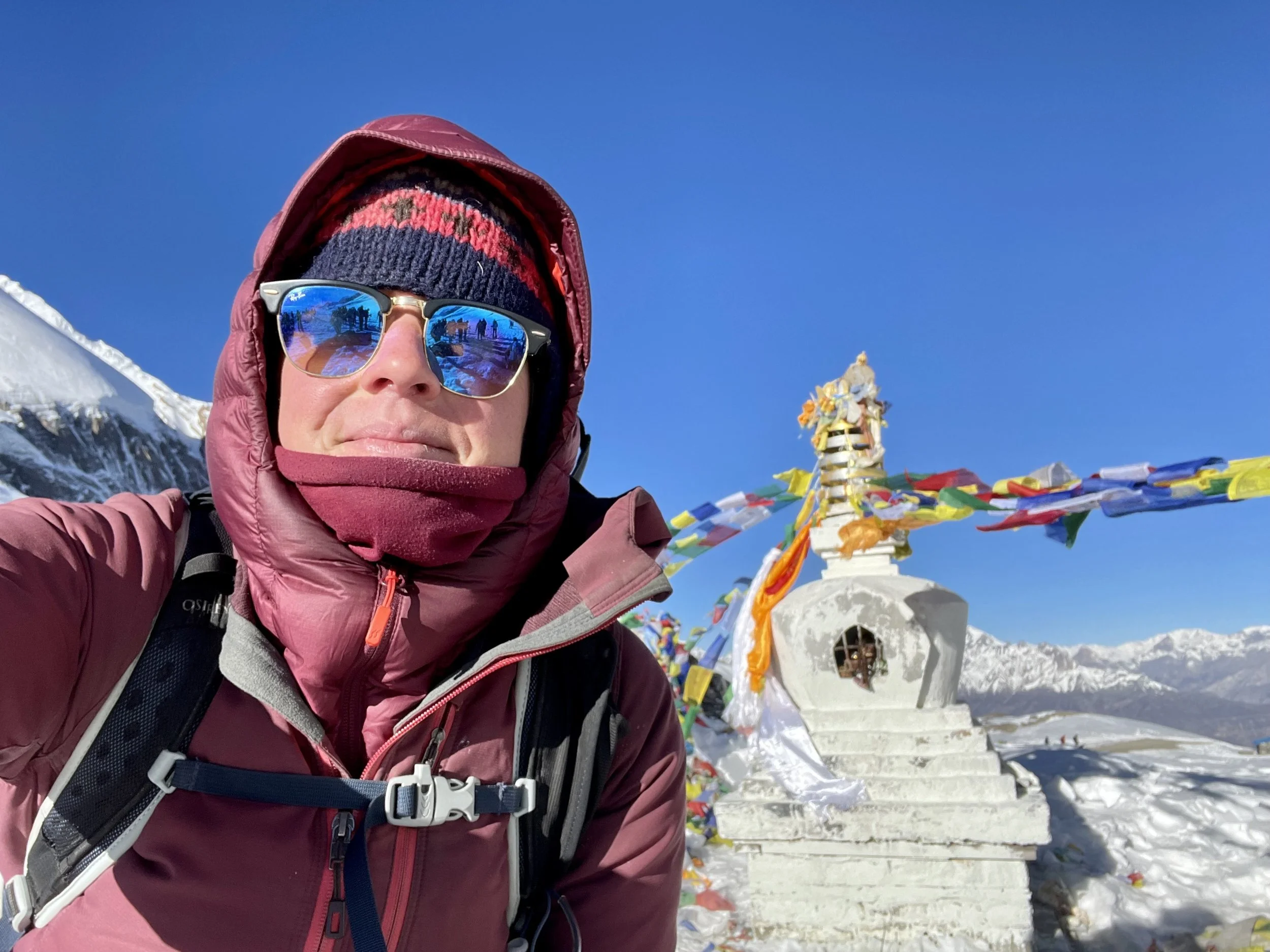Cat Goward wearing sunglasses, taking a selfie in a snowy mountain landscape Himalayas with prayer flags behind her.