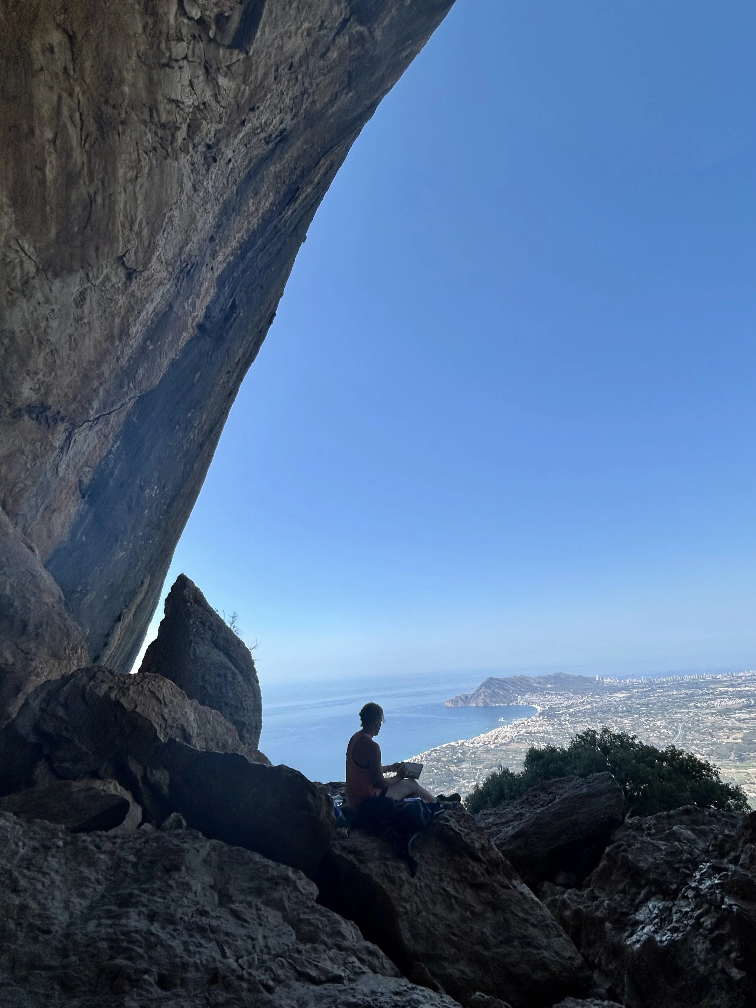 Cat Goward sitting on rocks in a cave overlooking a coastline and ocean, with a city in the distance.
